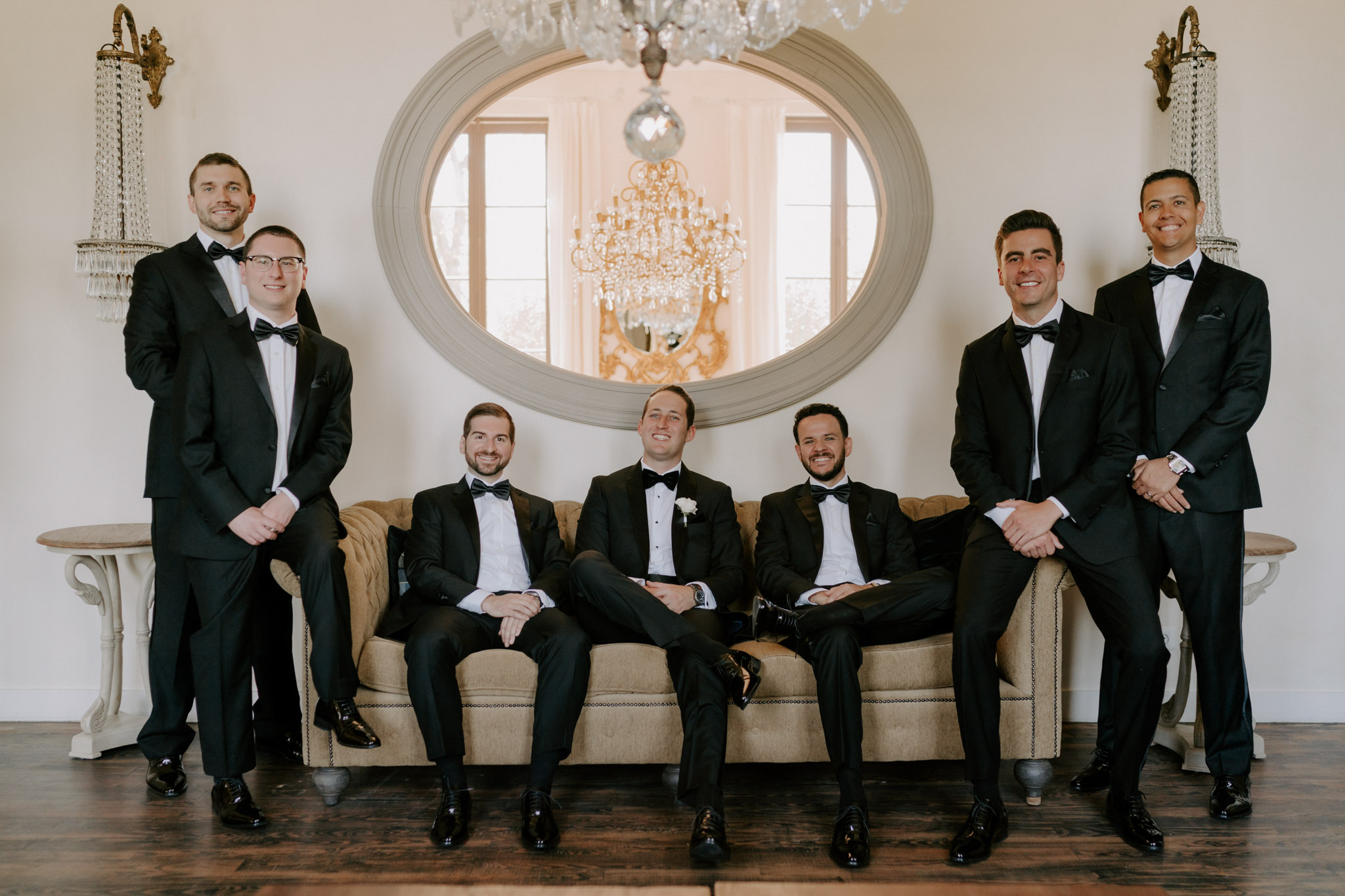 Groom and six groomsmen in black tuxedos posed on velvet sofa beneath baroque gilt mirror and sconces