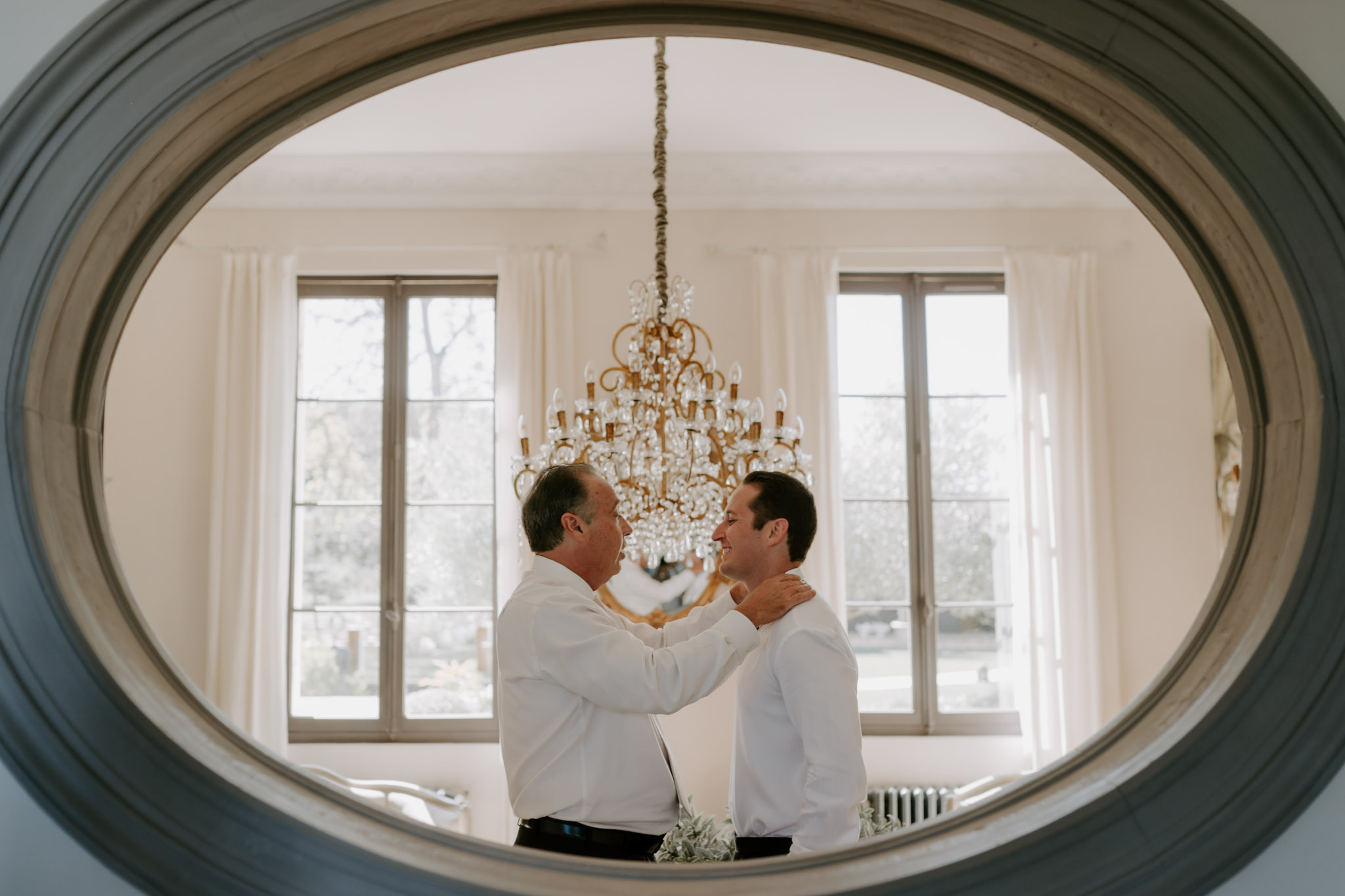 Father and groom reflected in oval mirror during getting ready with chandelier backdrop