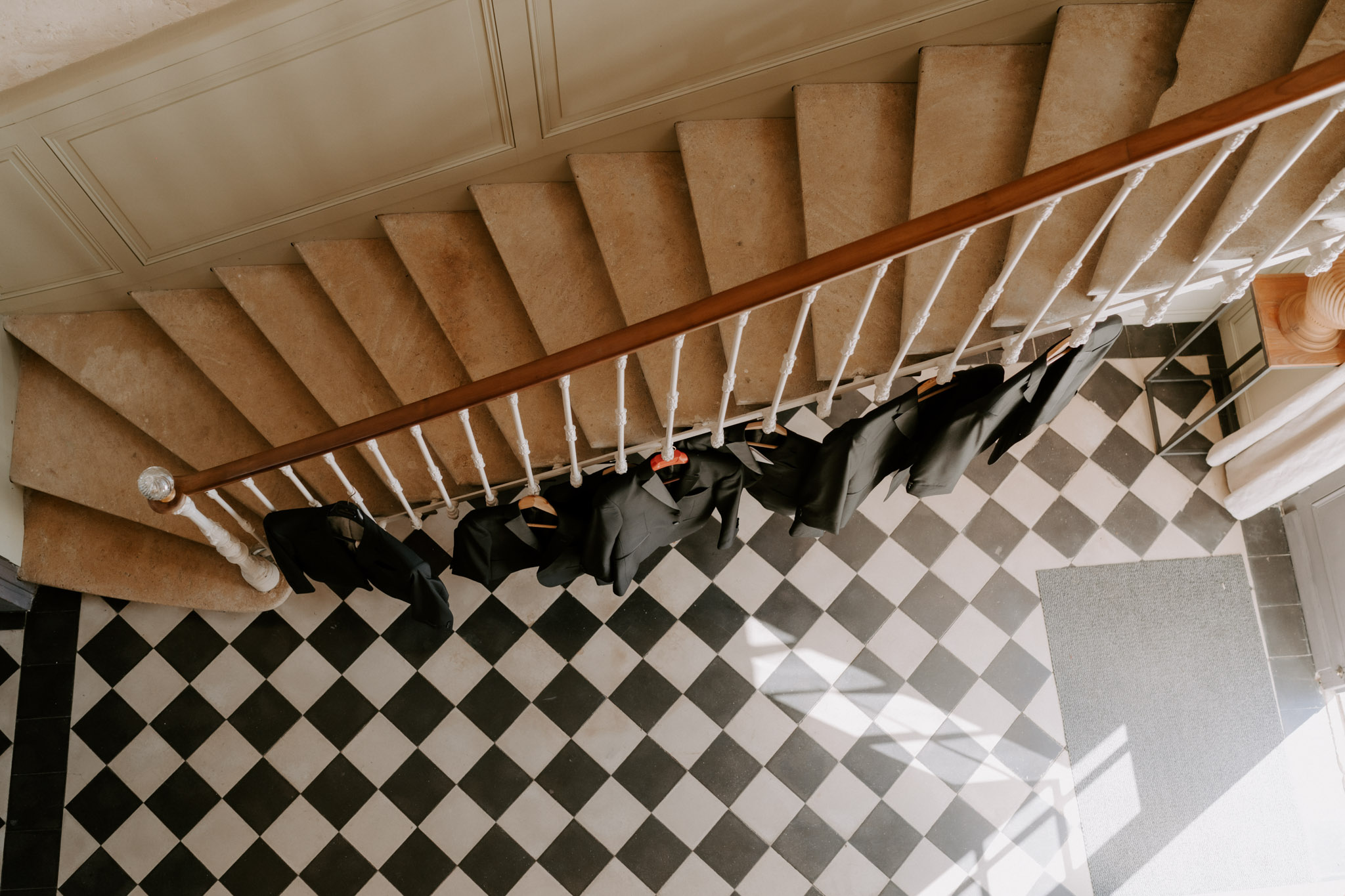 A getting-ready detail shot taken from an overhead perspective, showing approximately five to six dark navy or black suit jackets hanging on wooden hangers from the white wrought-iron balustrade of an interior staircase. The staircase features stone or marble steps and a warm wood handrail with white spindles. The ground floor below displays a classic black-and-white checkered tile floor, characteristic of a French château or manor house interior. No people are visible; the image focuses entirely on the groomswear arranged along the staircase railing. Wide overhead shot composition.