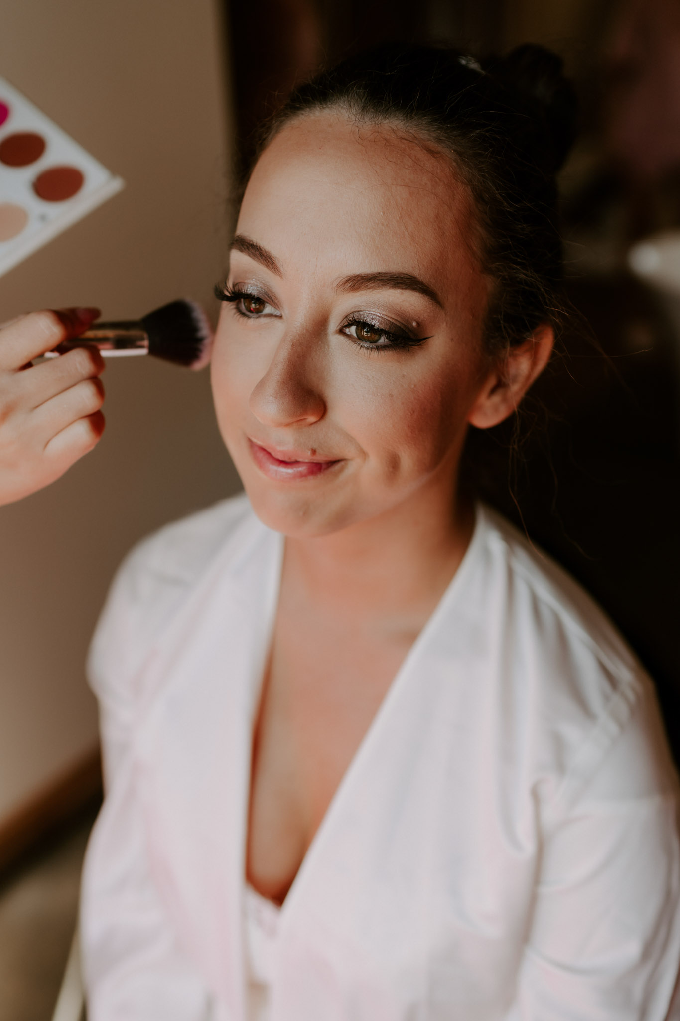 Bride in white satin robe having makeup applied with a powder brush during getting ready
