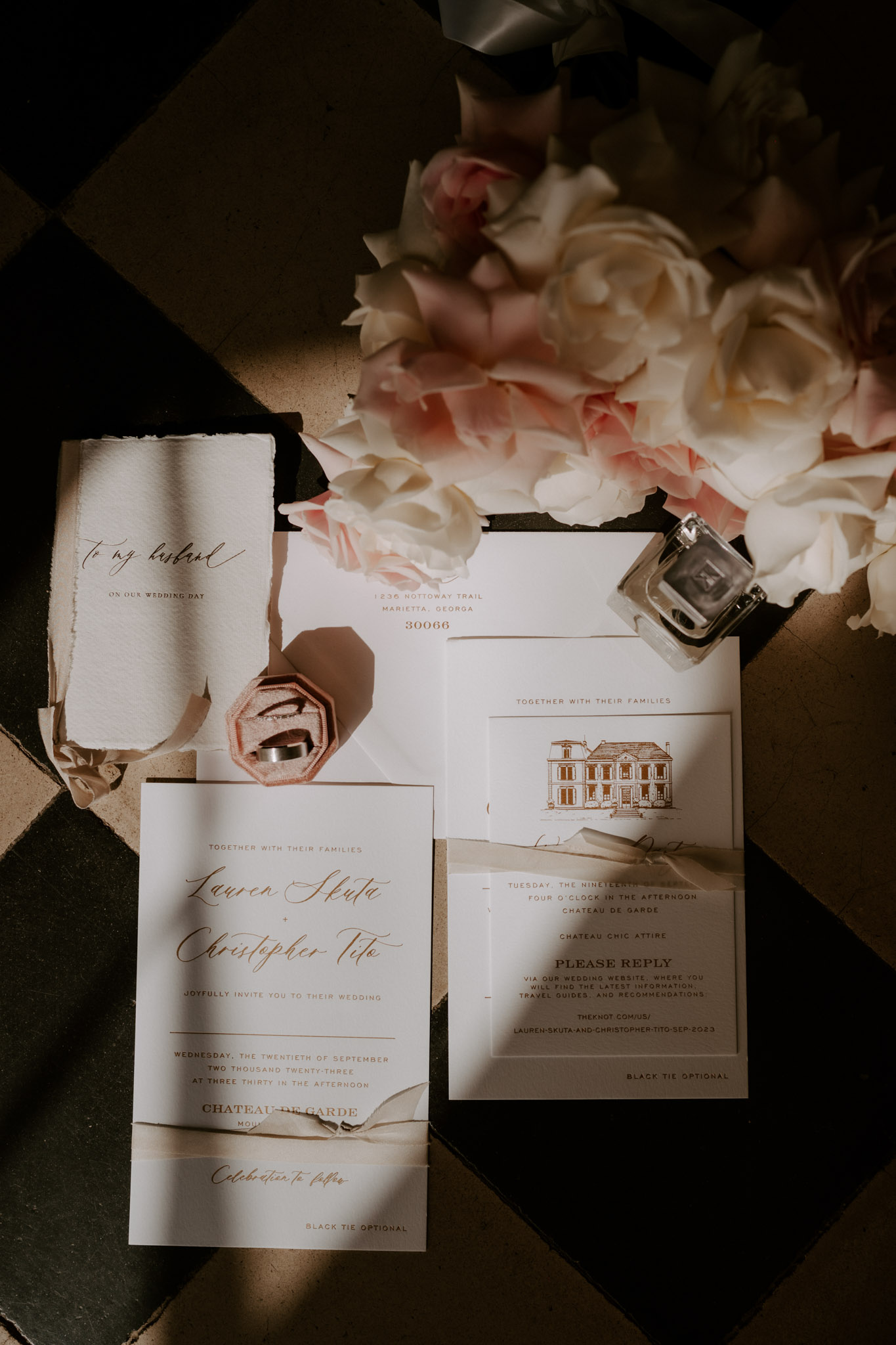 A flat lay detail shot of a wedding stationery suite arranged on a black and white checkered tile floor. The suite includes a main invitation card in ivory with gold/copper script calligraphy for Lauren Skuta and Christopher Tito at Chateau de Garde, a reply card featuring a hand-illustrated line drawing of the château, a vellum envelope, and a handwritten note reading 'To my husband on our wedding day' on deckle-edged paper. A blush pink velvet octagonal ring box holds two wedding bands, a small square glass perfume bottle sits nearby, and the arrangement is styled with blush pink and cream garden roses and peonies in soft focus in the upper portion of the frame. The overall stationery palette is ivory, gold, and blush, tied with a champagne satin ribbon, giving the suite a classic, formal aesthetic.
