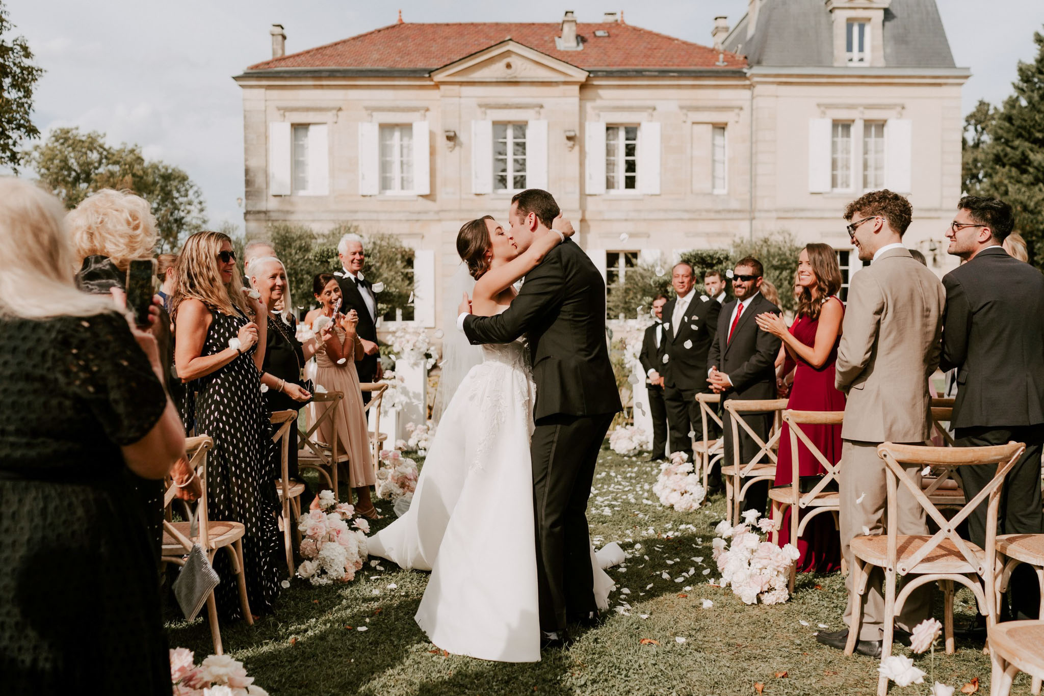 The bride and groom share their first kiss during an outdoor ceremony recessional on the lawn of a French château with a terracotta-tiled roof and stone façade. The bride wears a strapless white ballgown with lace detailing and the groom wears a black tuxedo; approximately 20 guests line the aisle on both sides, several tossing white flower petals. The aisle is lined with clusters of blush pink and white floral arrangements, and natural wood cross-back chairs are arranged on either side; guests are dressed in a mix of formal attire including black tie, a deep red gown, and a tan suit. The shot is a wide mid-distance image capturing the couple in the center of the frame with the château prominently featured in the background, giving it a classic, formal outdoor ceremony aesthetic. Potential venue feature image.