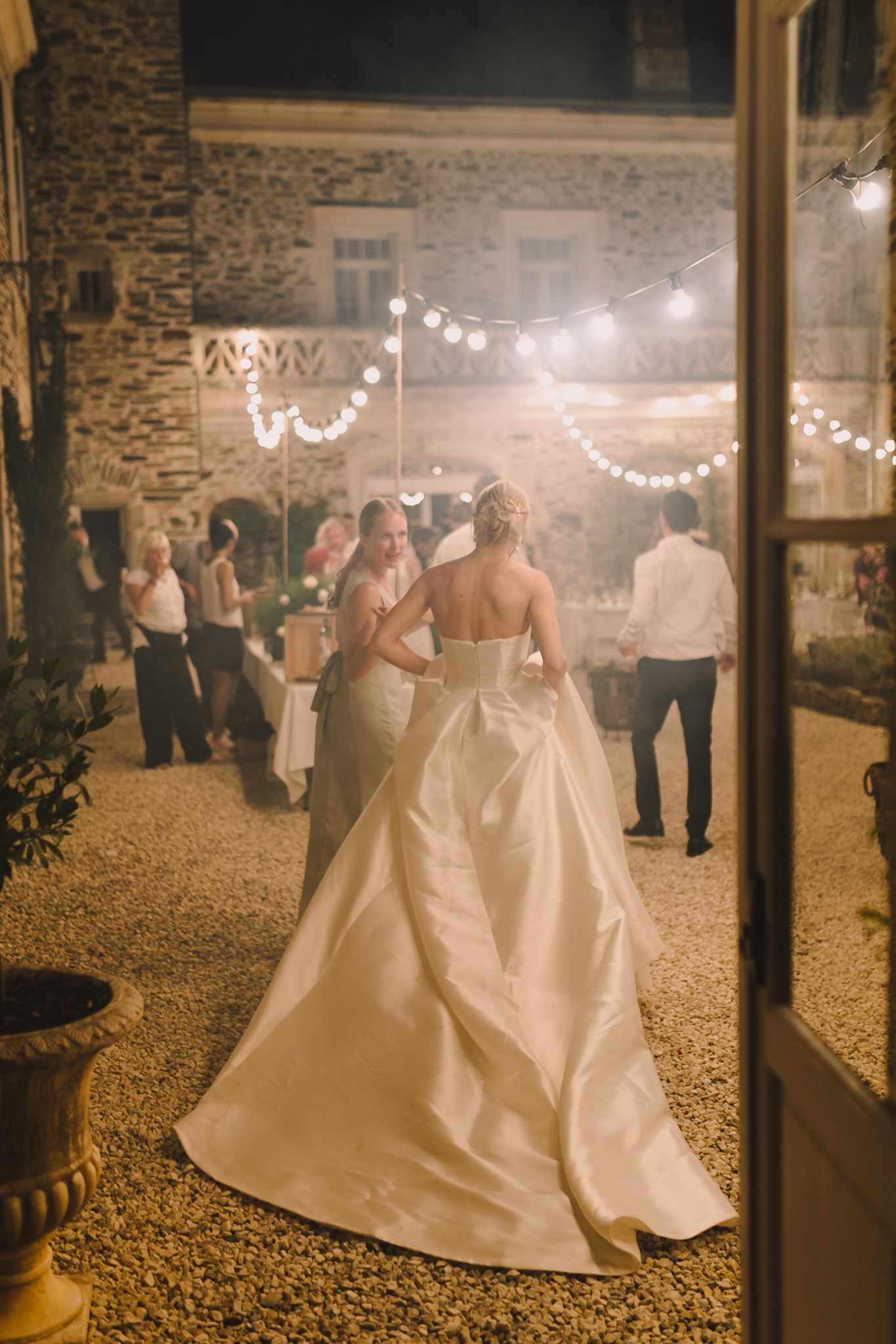 Bride in strapless ballgown walking into courtyard reception with string lights and guests from doorway