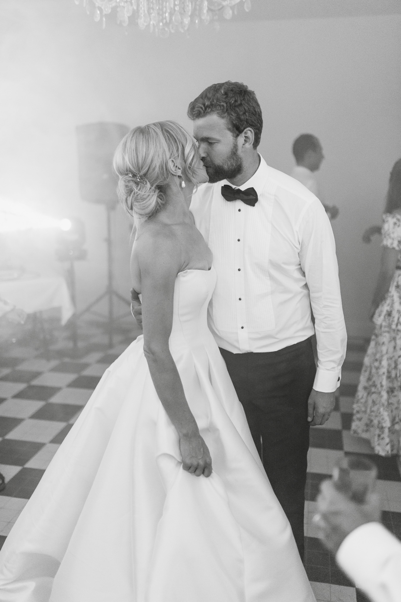 This black-and-white image captures the bride and groom sharing a kiss on the dance floor during what appears to be their first dance at an indoor reception. The bride wears a strapless ballgown with a full skirt and has her blonde hair styled in a low updo with a small hair accessory and drop earrings; the groom is dressed in a white tuxedo shirt with a black bow tie and dark trousers, with his jacket removed. The setting features a classic black-and-white checkered floor and a crystal chandelier visible in the upper background, suggesting a formal château or manor ballroom. A few guests are visible out of focus in the background, and the overall contrast is soft with bright highlights from event lighting on the left side of the frame — the shot is a mid-length portrait taken from slightly behind the bride.