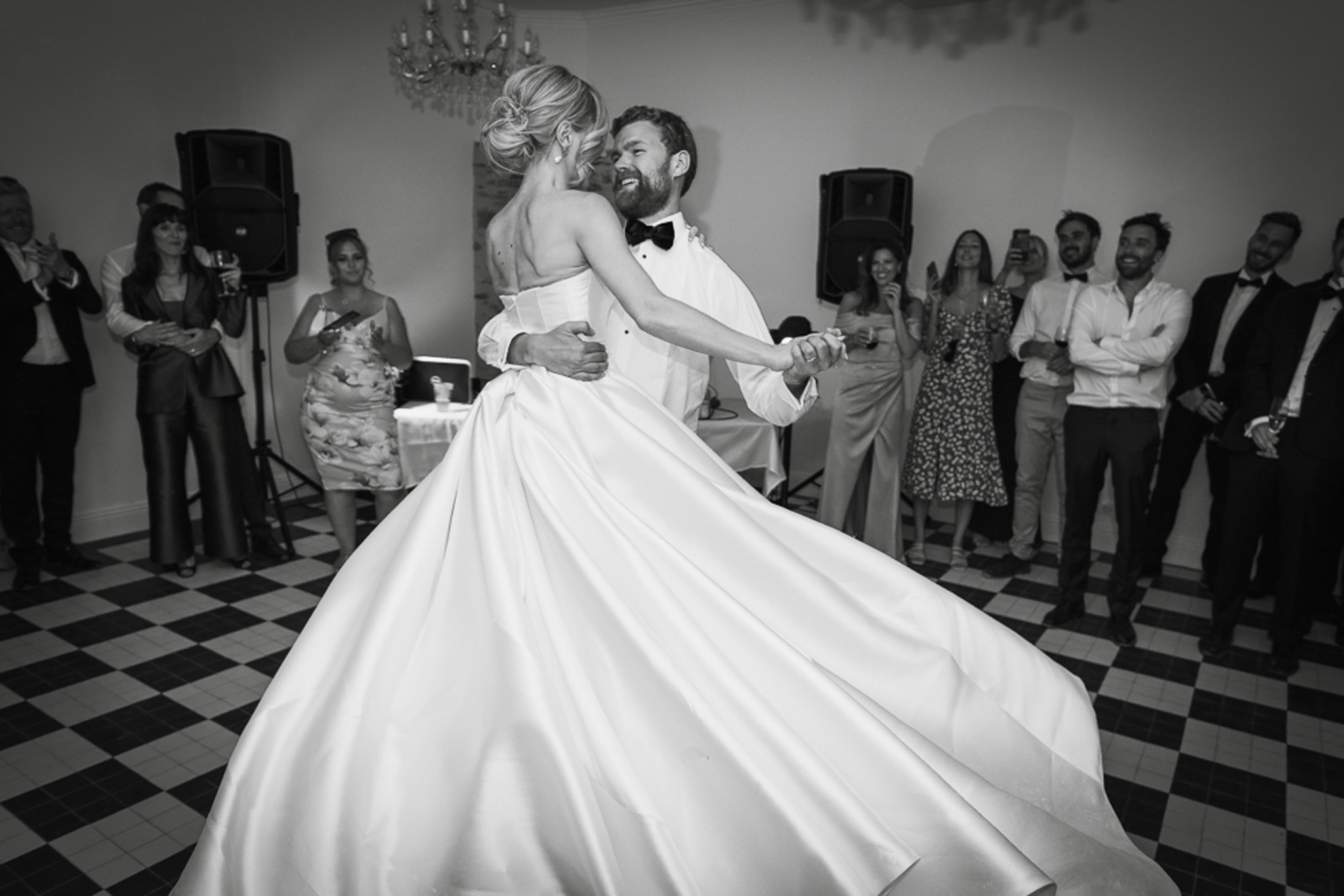 This black-and-white image captures the couple's first dance at an indoor reception. The groom, wearing a white dinner jacket with a black bow tie, lifts and spins the bride, whose full-skirted satin ballgown fans out dramatically around them. The bride wears her hair in an updo and the skirt's motion creates strong contrast against the black-and-white checkered tile floor. A crystal chandelier is visible overhead, reinforcing the classic interior setting. Approximately 12–15 guests line the perimeter of the dance floor, clapping and smiling, with PA speakers and a DJ laptop table visible in the background. The image is shot at mid-level with a wide-angle perspective that emphasizes the movement of the dress and the groom's expression of joy.