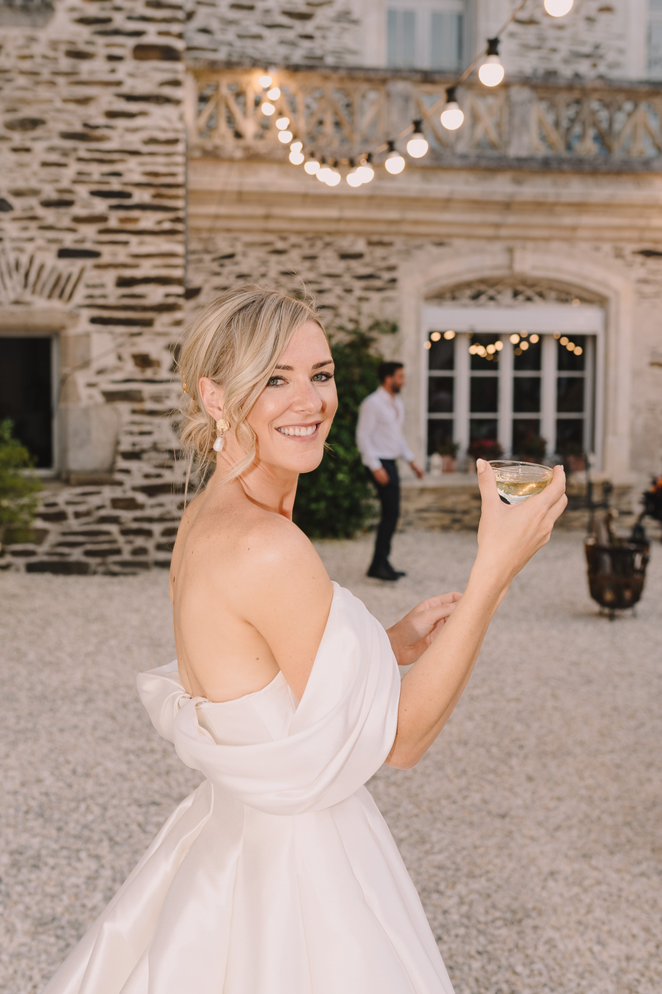 Bride in off-the-shoulder white ballgown holding coupe glass in chateau courtyard with string lights above