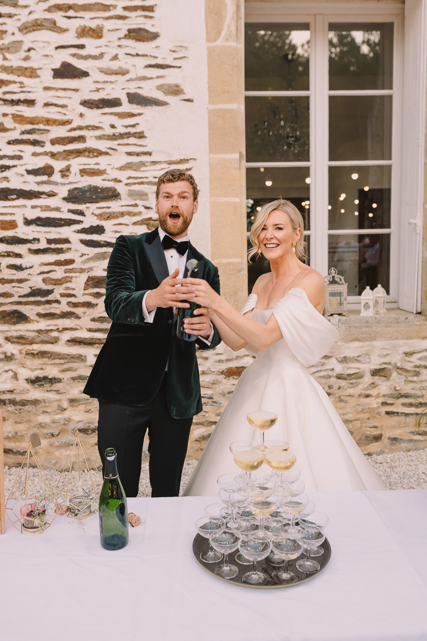 Groom in green velvet tuxedo and bride in white ballgown pour champagne tower at chateau terrace