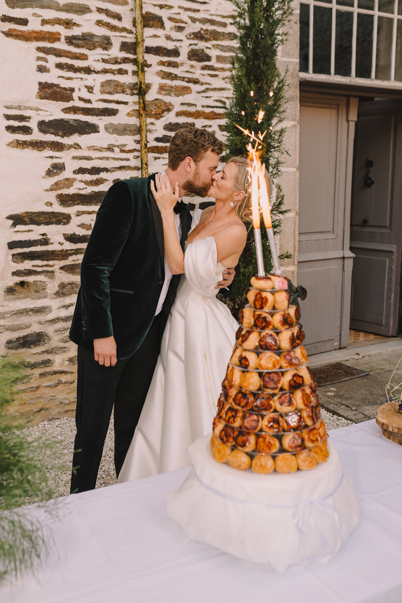 Couple kissing beside sparkler-topped croquembouche, groom in green velvet jacket, at rustic stone doorway