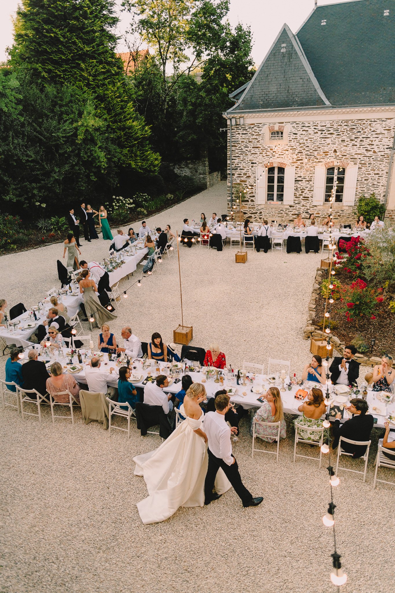 An outdoor wedding reception dinner is underway in a gravel courtyard in front of a traditional French stone manor house with a slate-grey pitched roof and white-shuttered windows. Long white-clothed rectangular tables are arranged in a curved horseshoe configuration seating approximately 50–60 guests, with white folding chairs and candles providing warm ambient lighting along the tables; festoon globe string lights are strung on wooden post stands across the space. The bride, wearing a white off-shoulder ballgown with a full skirt and train, and the groom in a white dress shirt and black trousers are walking together through the open center of the horseshoe arrangement, appearing to greet guests. Guests are dressed in a mix of formal attire including black tuxedos, teal and emerald gowns, and colorful cocktail dresses, and the overall styling is relaxed garden-party meets classic French country. The shot is taken from an elevated angle above and behind the couple, giving a wide overhead perspective of the full table layout and venue courtyard. Potential venue feature image.