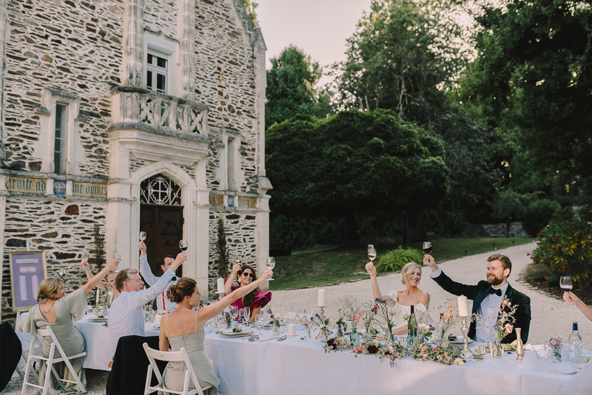 Wedding toast at outdoor reception table before stone chateau with guests raising glasses and wildflower centerpieces
