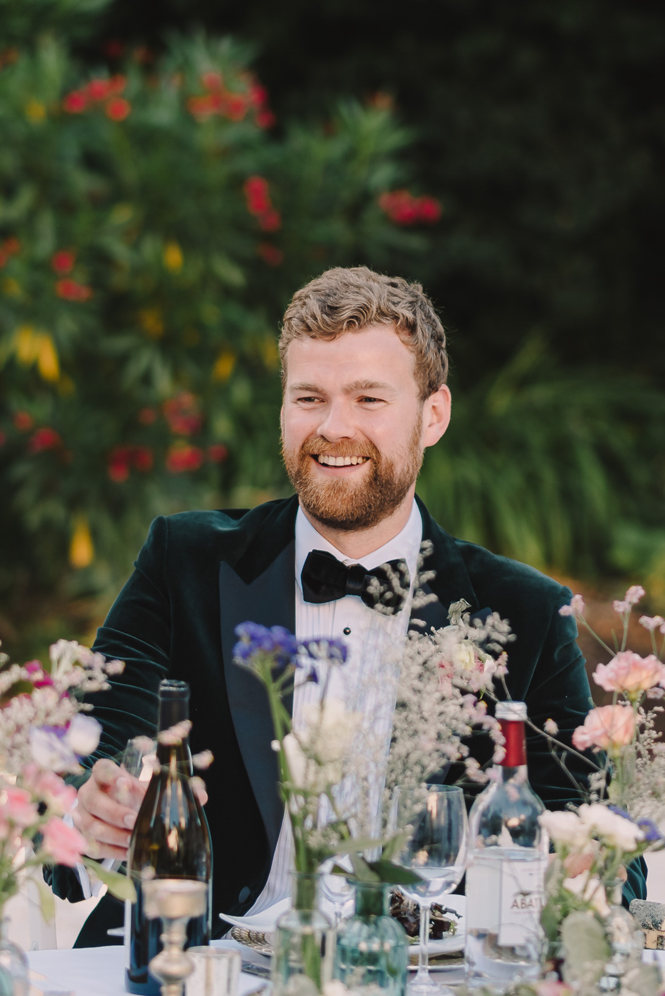 Groom in green velvet tuxedo laughing at dinner table with wildflower arrangements of sweet peas and irises