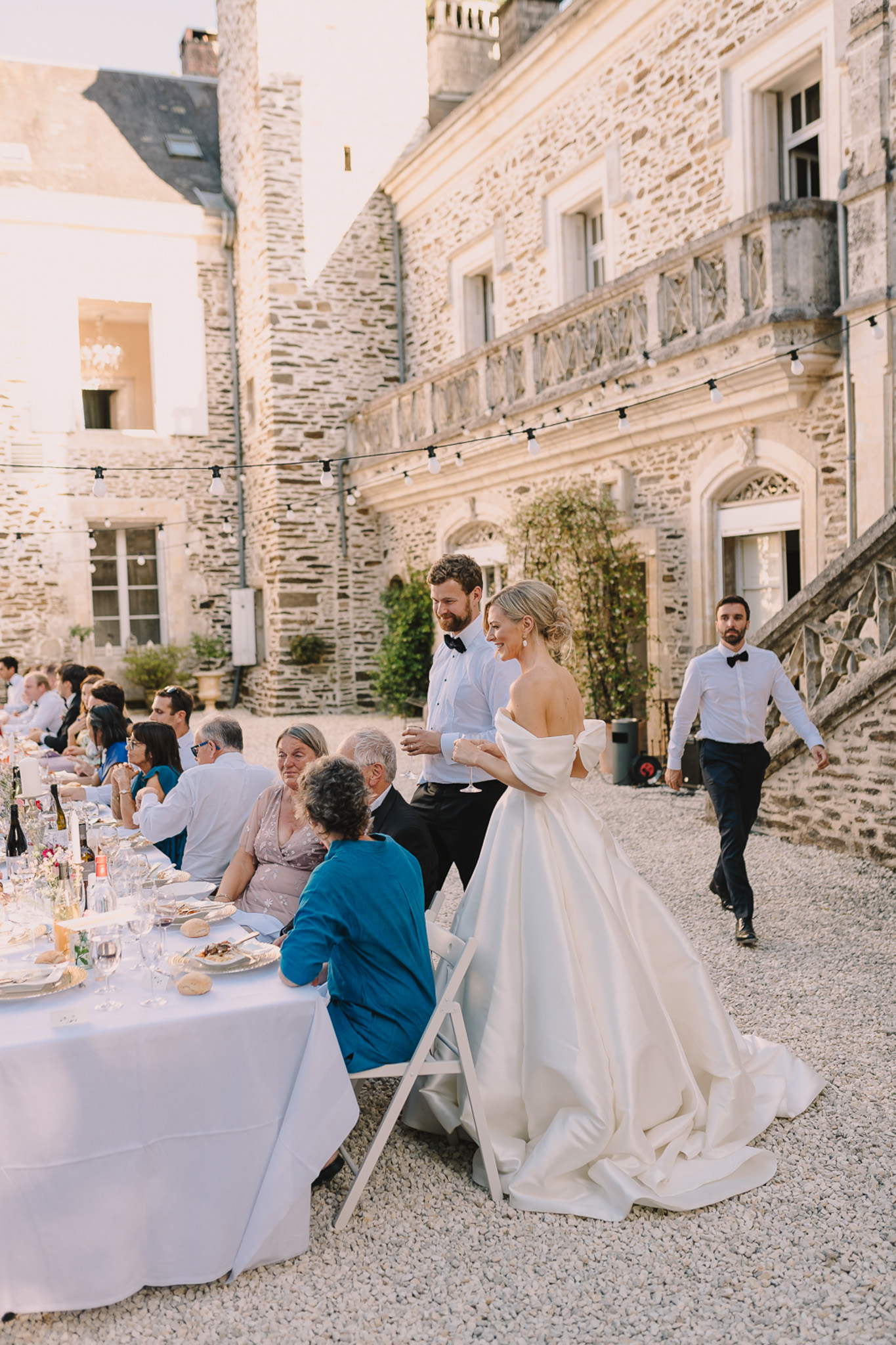 The couple is moving along the dinner reception tables in the courtyard of a French château, greeting seated guests during the outdoor evening meal. The bride wears an off-the-shoulder ivory ballgown with a full skirt and a dramatic train pooling on the gravel, while the groom is dressed in a black tuxedo with bow tie. A long banquet table dressed in a white linen cloth is set with glassware, candles, wine bottles, and plated food, with approximately 15–20 guests visible seated along it. Globe string lights are strung across the courtyard between the château's historic stone façade walls, adding warm ambient lighting as the golden hour light falls across the scene. A second man in a white shirt and bow tie approaches in the background near the château's ornate exterior staircase. The composition is a medium-wide candid shot capturing the couple mid-movement alongside the table. Potential venue feature image.