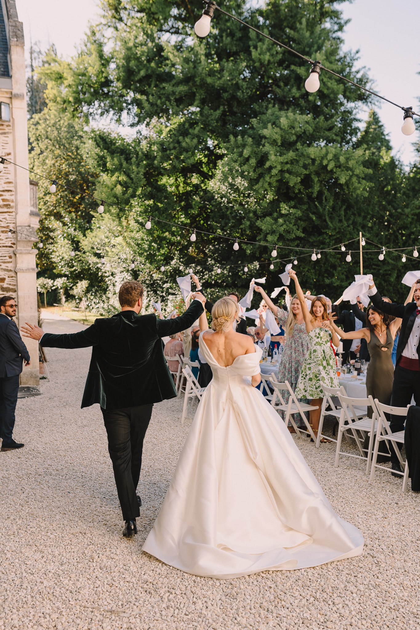Bride and groom make their grand entrance as guests wave white napkins at an outdoor chateau courtyard reception