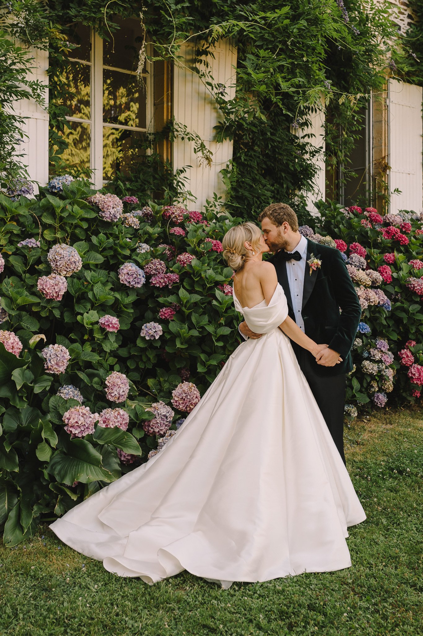 A couple portrait taken outdoors in a garden setting, with the bride and groom kissing in front of a large hydrangea border in bloom with pink, mauve, blue, and red flower heads. The bride wears an ivory off-the-shoulder ball gown with a dramatic full skirt and a low open back, with her hair styled in a low chignon. The groom wears a forest green velvet tuxedo jacket with a black bow tie and a small floral boutonniere. The white-shuttered building facade behind them is covered in climbing vines and wisteria. The shot is a medium full-length portrait framing both figures from head to toe, with the bride's gown train spreading across the lawn.