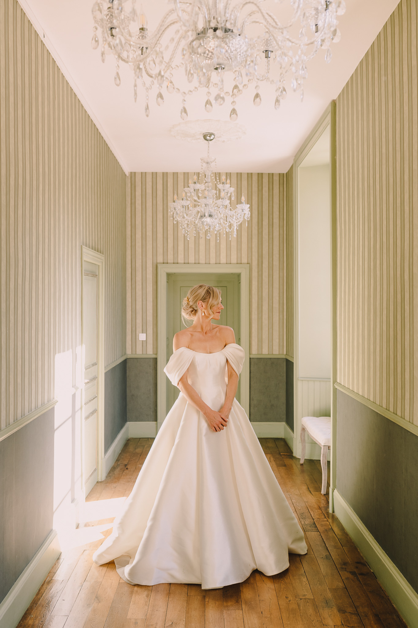 A bridal portrait taken indoors in a hallway or antechamber of what appears to be a French château. The bride stands alone, turned slightly to one side with her hands clasped in front, wearing an ivory off-the-shoulder ball gown with a full satin skirt and draped sleeve details. Her blonde hair is styled in a low updo and she wears statement drop earrings. The room features vertical striped wallpaper in sage green and cream tones, sage-painted wainscoting, wide-plank hardwood floors, and two crystal chandeliers hanging from a white ceiling. Natural light streams in from the left, casting a soft shadow across the floor. The composition is a full-length portrait shot taken from a low-to-mid angle, centering the bride beneath the chandeliers with a doorway visible behind her. Potential venue feature image.