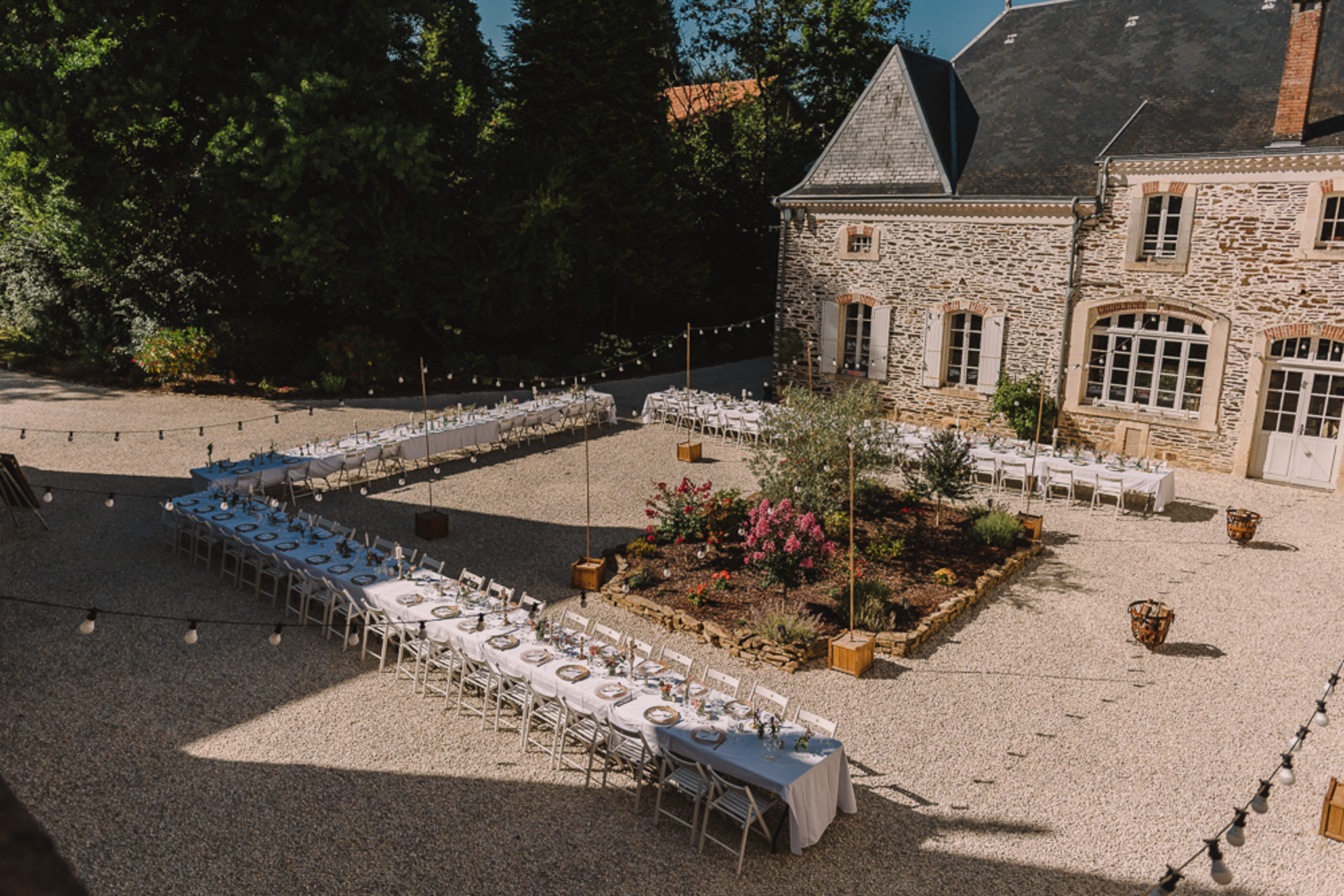 Aerial reception setup L-shaped banquet tables white and blue linen festoon lights on gold poles at stone manor courtyard