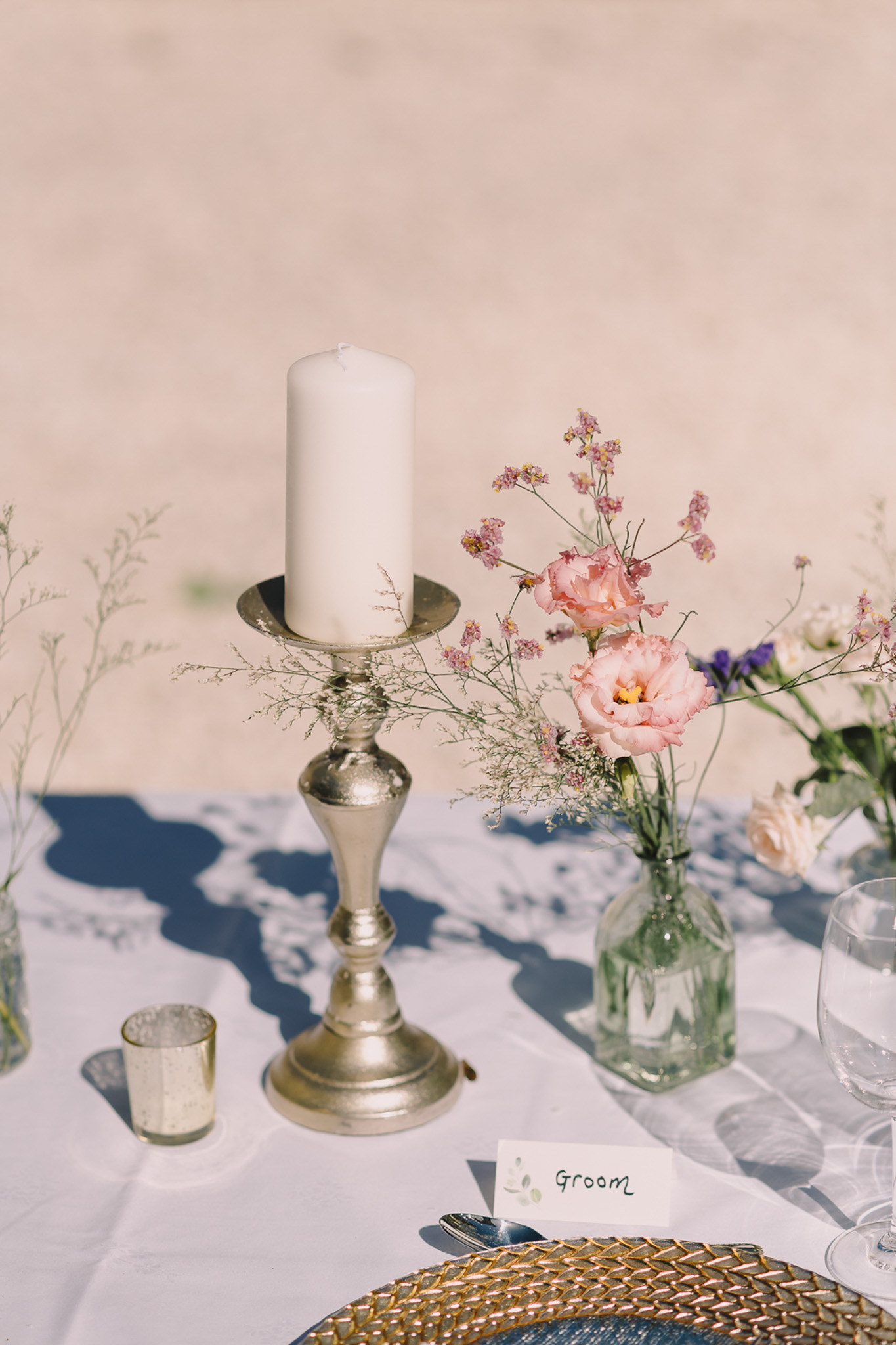 Reception place setting with silver candlestick, blush lisianthus bud vases, gold charger, and groom place card