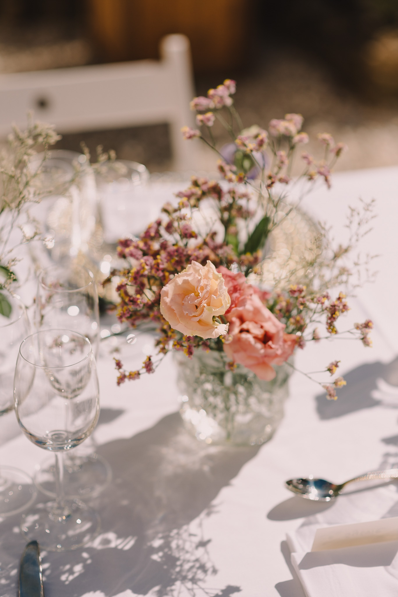 A close-up detail shot of an outdoor wedding reception table set in bright natural light. The centerpiece is a clear glass vase containing a loosely arranged wildflower-style bouquet with peach and coral roses, dusty mauve limonium, white gypsophila, and small filler greenery. The white linen tablecloth is dressed with clear wine glasses, silver cutlery including a spoon and fork, and a cream-colored taper candle partially visible at the edge. A white wooden chair is softly blurred in the background. The overall decor palette is soft and romantic with a relaxed, garden-inspired aesthetic.