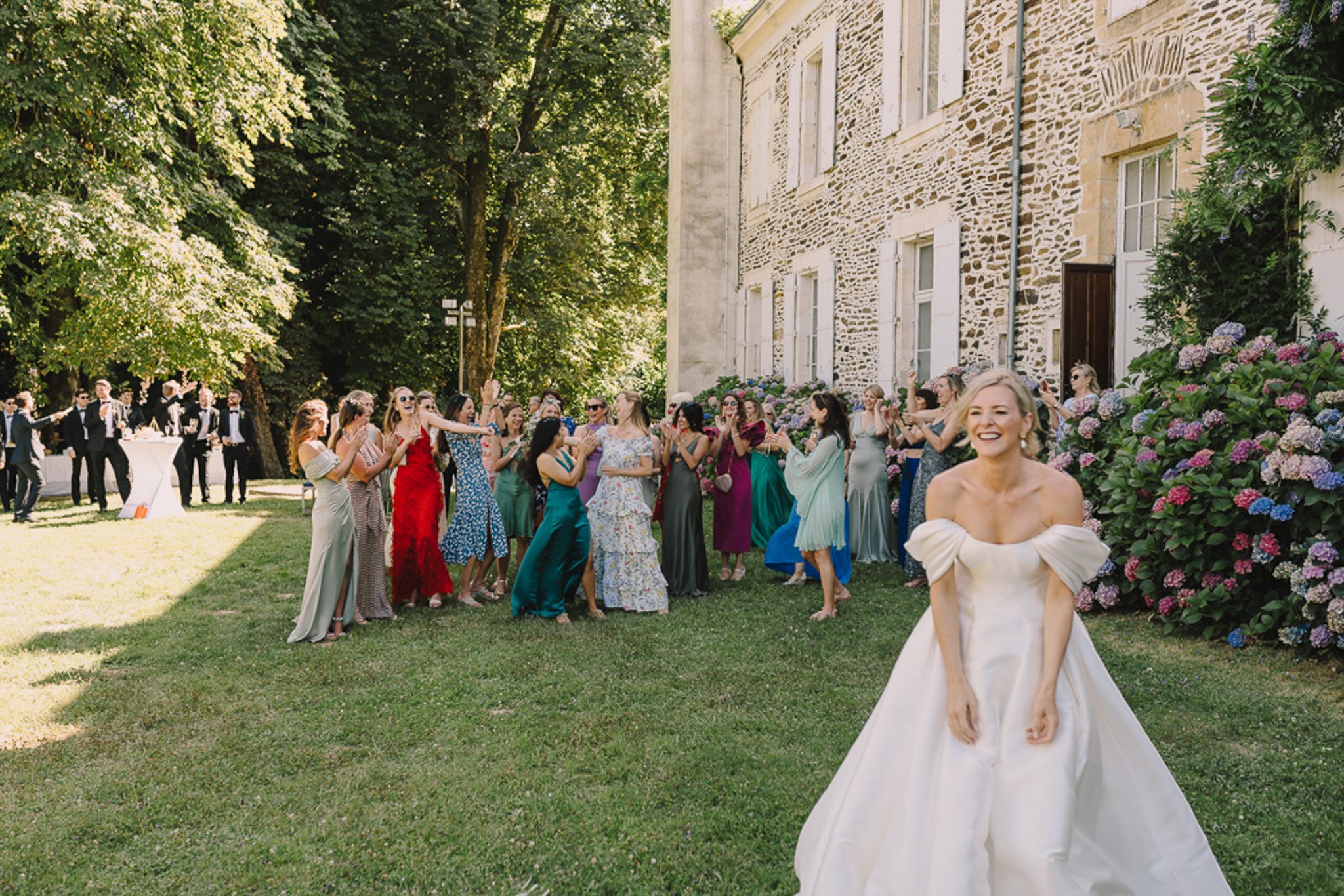 The bride is captured mid-bouquet toss on the lawn of a French château during what appears to be a cocktail hour or reception. She is in the right foreground, wearing an off-the-shoulder white ball gown with structured sleeves, laughing as she faces away from a large crowd of approximately 25–30 female guests who are reaching and reacting excitedly behind her. The guests are dressed in a vivid mix of colorful formal attire including red ruffled, teal satin, sage, burgundy, blue, and floral patterned gowns and dresses. In the background to the left, a smaller group of male guests in black tuxedos stands near a cocktail table. The setting is outdoor on a manicured lawn beside a stone château building with white-shuttered windows, with large blooming pink and blue hydrangea bushes visible against the building's facade. The shot is taken from a wide-angle perspective at ground level, capturing both the bride's joyful expression and the full crowd reaction in a single frame.