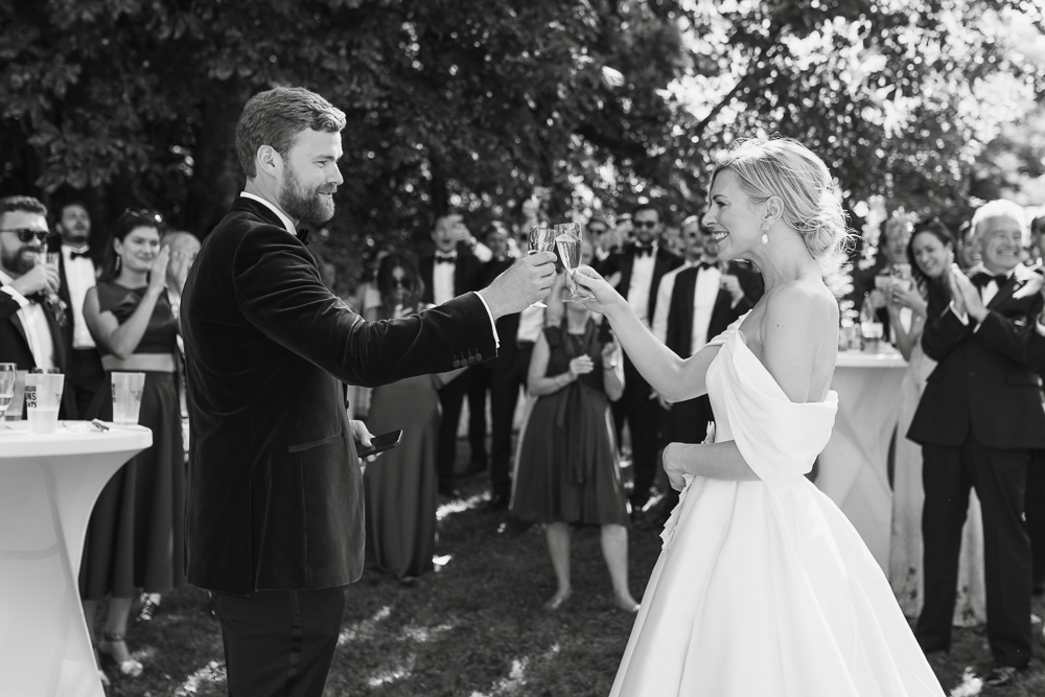 Black-and-white photo of bride and groom toasting champagne flutes surrounded by applauding guests at outdoor cocktail hour