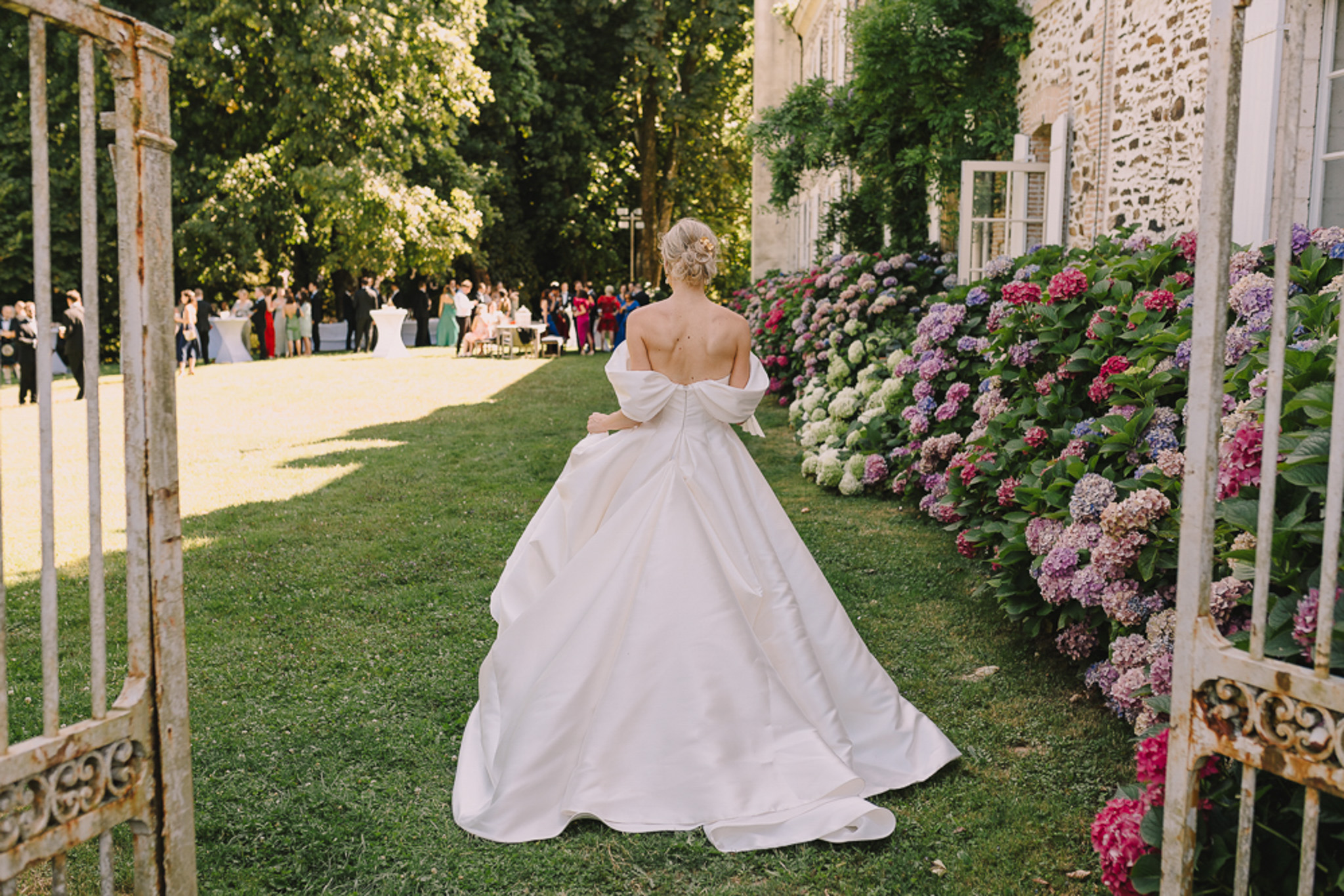 Bride in off-shoulder ball gown walking through iron gates toward cocktail gathering at chateau with hydrangea borders