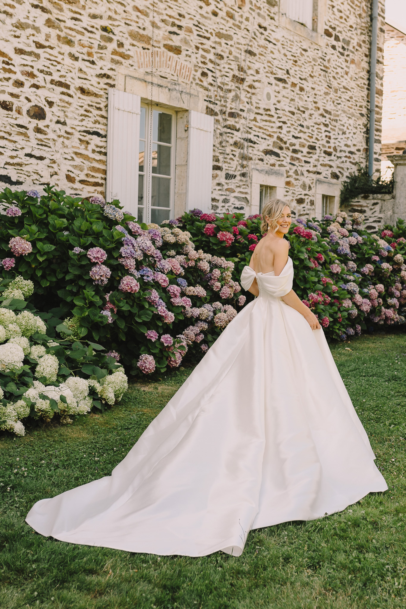 Bride glancing back in ivory bow-back ballgown with cathedral train spread before hydrangea border