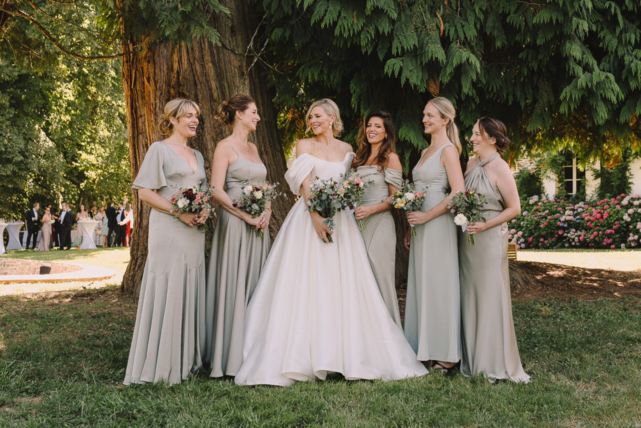 Bride with five bridesmaids in mismatched sage green dresses holding pink and burgundy bouquets by tree