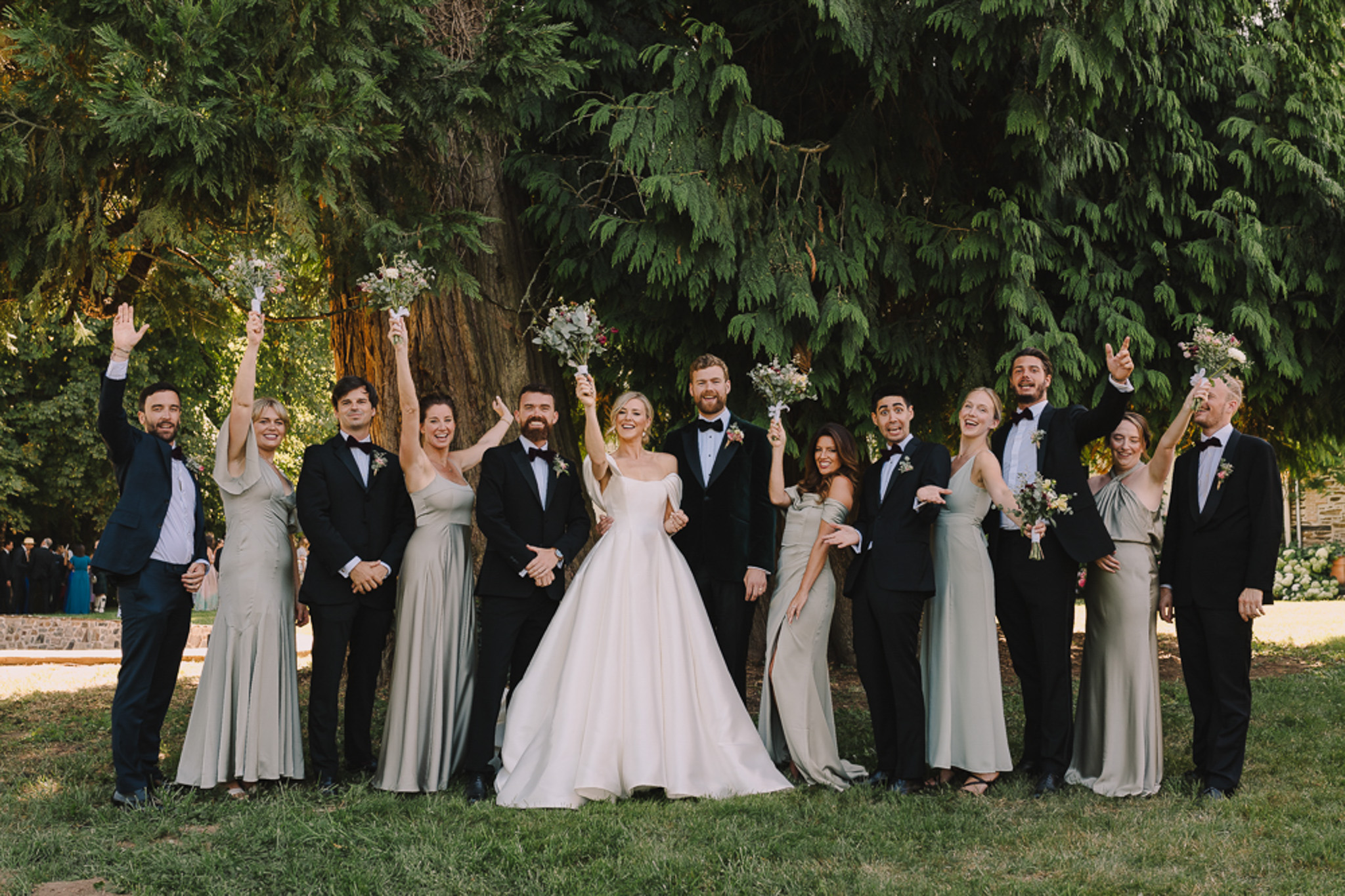 Bride, groom, and twelve attendants in sage gowns and tuxedos celebrate on chateau lawn
