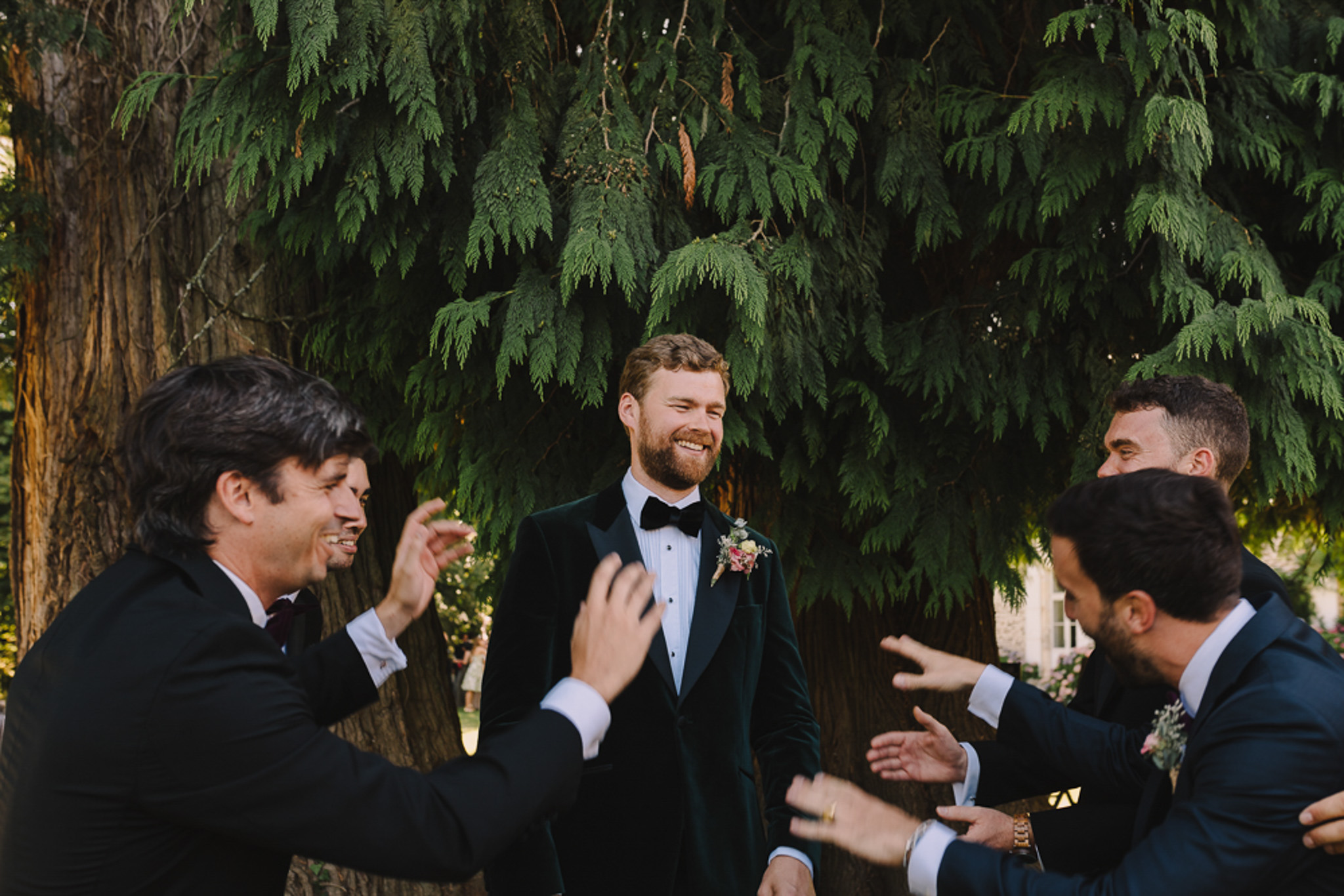 The groom stands outdoors surrounded by approximately four groomsmen or male guests who are clapping and gesturing toward him in celebration, likely during the cocktail hour or pre-ceremony gathering. The groom wears a deep forest green velvet tuxedo jacket with a black bow tie, white dress shirt, and a boutonniere featuring pink and cream flowers with greenery. The men around him wear dark navy and black suits, with one wearing a burgundy bow tie; at least one other man also wears a boutonniere. The shot is a mid-length portrait framed against a large conifer tree, captured in natural outdoor light with a candid, documentary feel.