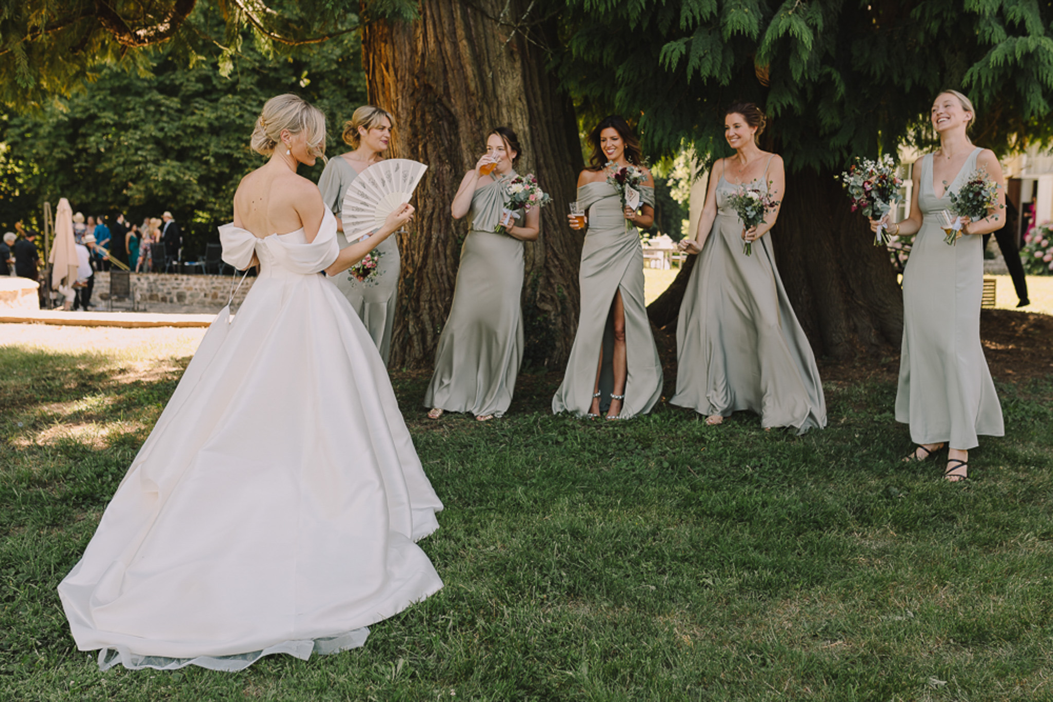 The bride stands facing five bridesmaids in an outdoor garden setting, photographed from behind in a wide shot. The bride wears an off-the-shoulder white ballgown with a large bow detail at the back and holds a white decorative hand fan; her blonde hair is styled in an updo. The five bridesmaids wear mismatched sage green satin floor-length gowns in varying silhouettes including off-the-shoulder, draped, and V-neck styles. Several bridesmaids hold bouquets featuring deep burgundy, pink, and white blooms with trailing greenery, while some also hold drinks. The group appears relaxed and candid, with laughter visible among the bridesmaids. In the background, wedding guests are visible gathered near what appears to be a stone terrace or low wall, suggesting this is during the cocktail hour.