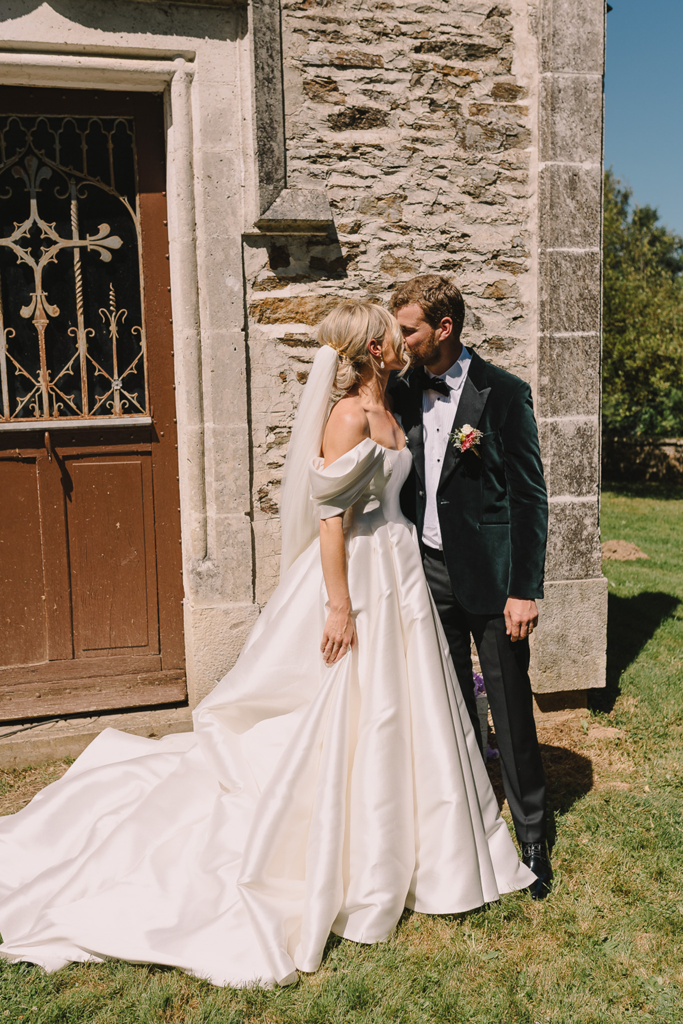A couple portrait taken outdoors beside a stone chapel or church building with a ornate wrought-iron and wood door. The bride wears an ivory off-the-shoulder satin ball gown with a long cathedral train and a sheer veil worn in a low updo; she has drop earrings. The groom wears a dark forest green velvet tuxedo jacket with black trousers, a white dress shirt, black bow tie, and a small mixed-color boutonniere. The two are kissing in profile. The composition is a mid-length portrait shot taken in natural daylight with strong shadows indicating bright midday sun.