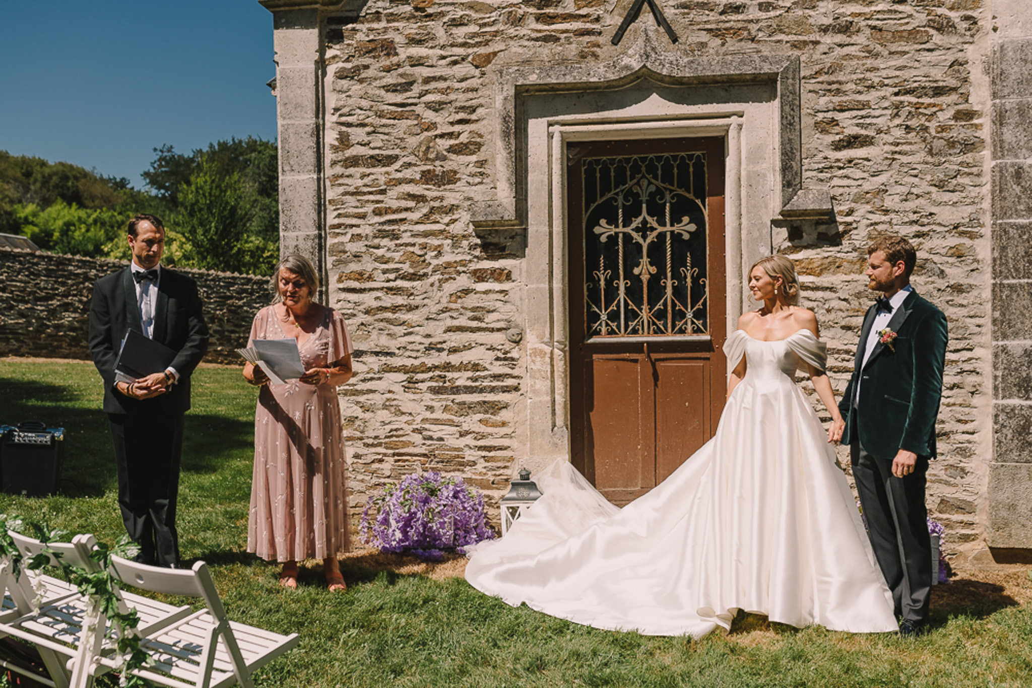 Outdoor ceremony before stone chapel with bride in ball gown, groom in green velvet blazer, and officiant
