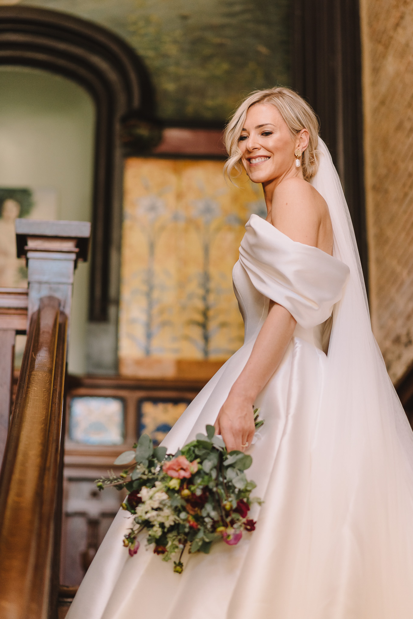 A bridal portrait of a single bride standing on an interior staircase, turning back toward the camera with a smile. She wears an ivory off-the-shoulder satin ball gown with a full skirt and a long cathedral-length veil, paired with pearl drop earrings and a loose blonde updo. She holds a loosely arranged bouquet featuring deep burgundy ranunculus, blush and coral open blooms, small white filler flowers, and trailing eucalyptus greenery. The interior setting features ornate painted wall panels in gold and blue-green tones, a large arched mirror, and a dark wood staircase banister, suggesting a historic château or manor house interior with a classic, formal aesthetic. The shot is a three-quarter portrait with the background softly blurred to draw focus to the bride.