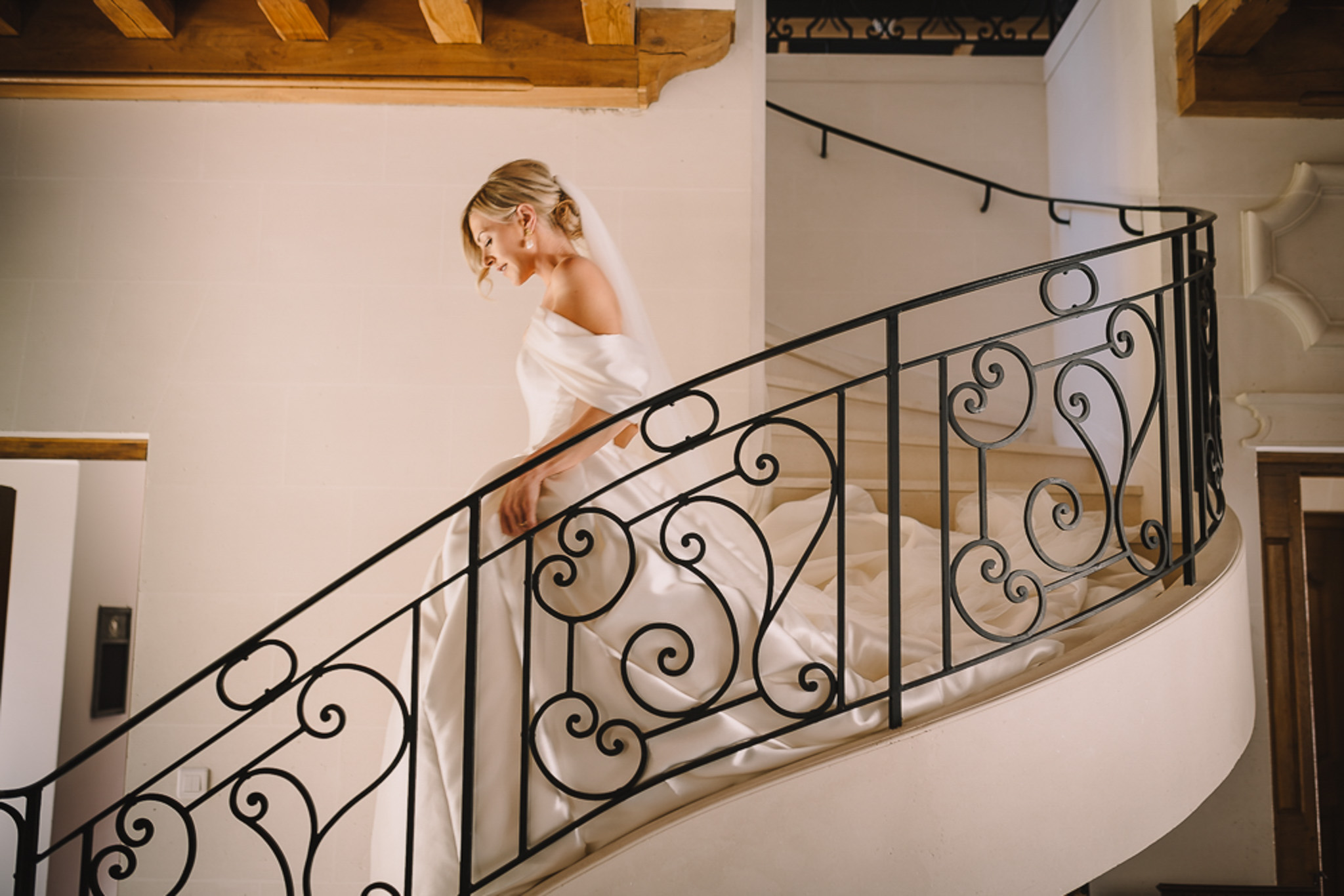 Bride in off-shoulder satin ballgown descends curved staircase with iron scrollwork and cathedral veil