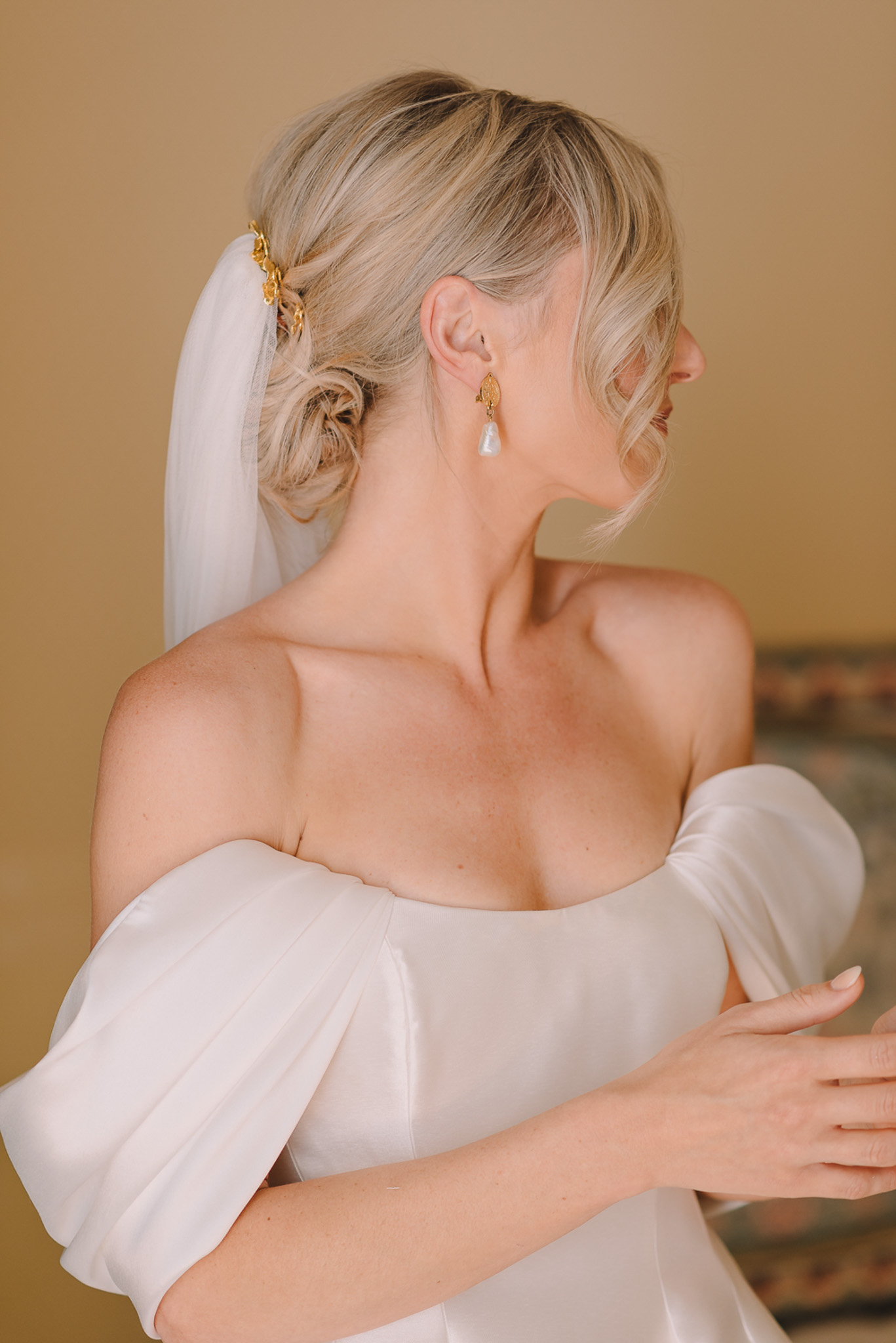 Close-up portrait of a bride photographed from a three-quarter rear angle, looking to the side against a warm beige wall. She wears an ivory off-the-shoulder gown with voluminous puff sleeves in a smooth satin fabric. Her blonde hair is styled in a low twisted updo with a white veil attached at the crown using small gold floral hair clips. She wears gold drop earrings featuring a leaf-shaped top element and a baroque pearl pendant. The overall styling palette is ivory, gold, and white, with a clean modern-classic aesthetic.