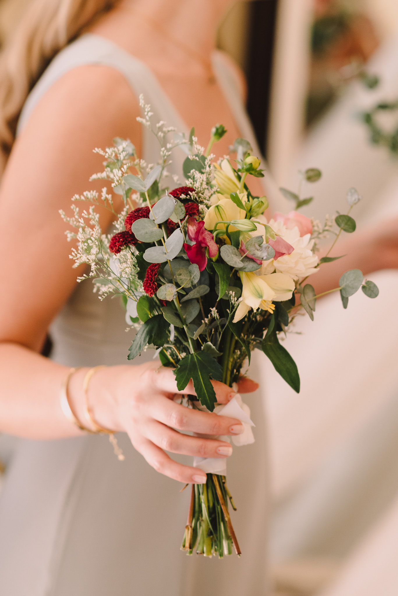 A close-up detail shot of a bridesmaid holding a small, loosely arranged bouquet with a white ribbon wrap around the stems. The bouquet includes deep burgundy celosia, soft yellow lilies, blush pink sweet peas, a cream dahlia, white astilbe or limonium, and silver dollar eucalyptus with assorted greenery, giving it a garden-style, slightly wildflower feel. The bridesmaid is wearing a light grey sleeveless dress and gold bangle bracelets. The background is softly blurred, revealing a second similarly dressed person in the background.