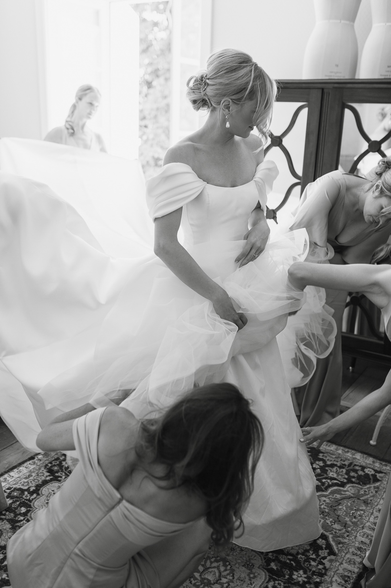 This black-and-white getting-ready shot captures a bride being assisted by at least two bridesmaids as they arrange the voluminous tulle skirt of her off-the-shoulder ball gown. The bride wears her hair in an updo with a decorative hair accessory and drop earrings, and the dress features structured off-the-shoulder sleeves with a full layered tulle skirt. The bridesmaids are dressed in light-toned spaghetti-strap gowns. The indoor setting appears to be a well-appointed dressing room with a patterned area rug, dark wood furniture, and a dress form visible in the background; a mirror in the background reflects a third figure, likely another bridesmaid or the bride's reflection. The image is shot from a slightly elevated angle, giving a dynamic, swirling quality to the full skirt as it is being fluffed and arranged. High contrast between the bright white gown and the darker surroundings gives the image strong tonal depth.