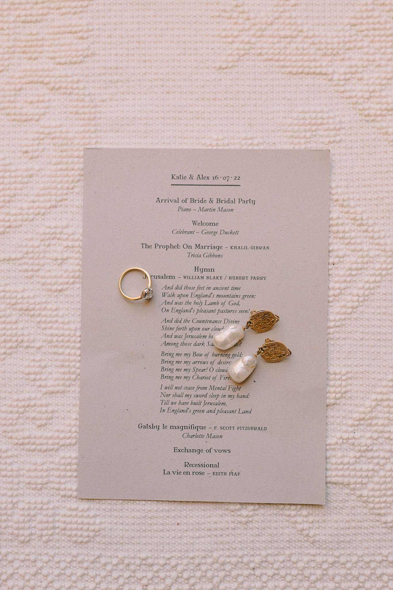 Ceremony program card with gold coin pearl earrings and diamond ring on grey paper and cream fabric