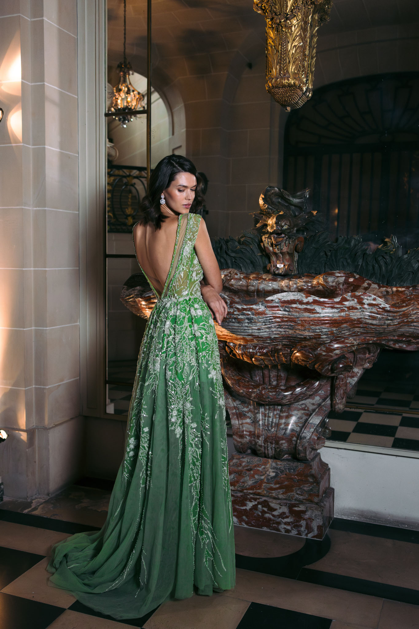 Woman in emerald green beaded gown with open back photographed from behind in ornate chateau interior