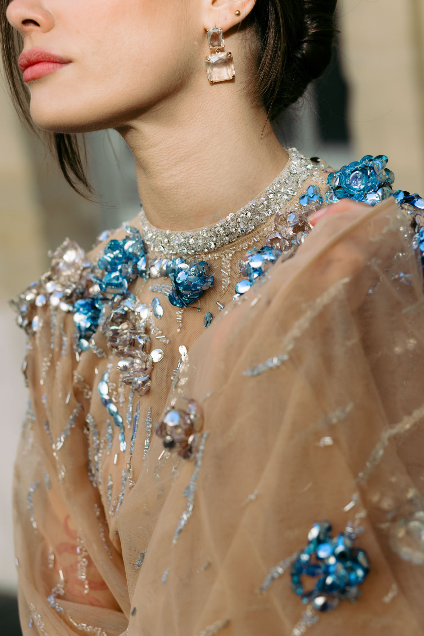 A close-up detail portrait of a woman, likely a bride, wearing a nude/champagne tulle dress heavily embellished with blue sequin rosette appliqués, silver bugle beads, and scattered mauve and clear crystal embellishments across the bodice and shoulders. The high neckline is trimmed with silver crystal beading. She accessorizes with gold drop earrings featuring clear faceted rectangular stones in a two-tier design, and her dark hair is styled in a low updo. The composition is a tight detail shot cropped from chin to mid-torso, with the background blurred, drawing full focus to the dress embellishment and jewelry. The styling aesthetic is formal and ornate, with a modern couture feel.