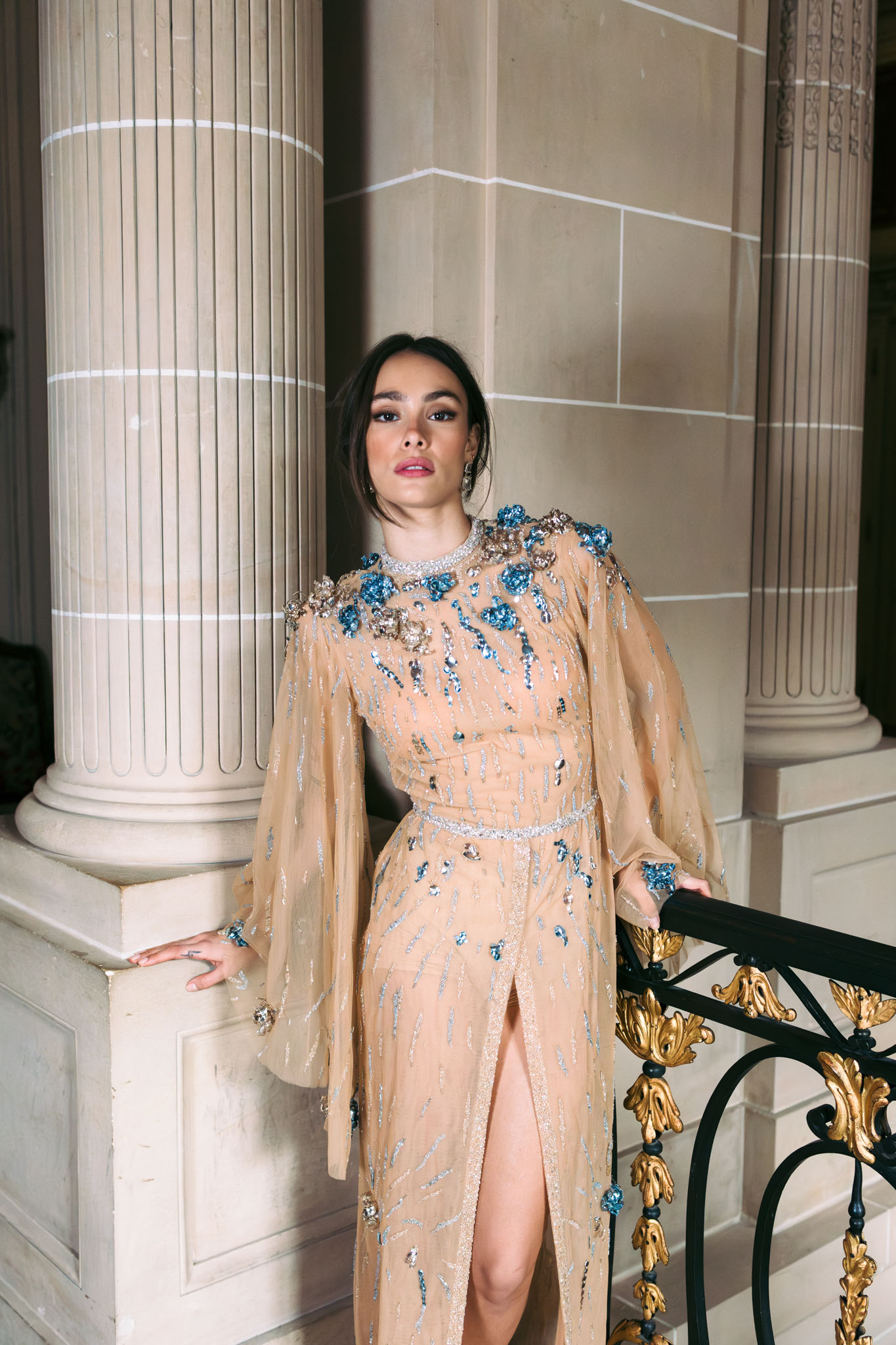 A portrait of a woman, likely the bride or a guest, posing between classical stone columns with a ornate black and gold wrought-iron railing visible to the right. She is wearing a floor-length champagne/nude sheer gown with long cape-style sleeves, heavily embellished with blue crystal floral appliqués at the shoulders, a crystal-encrusted neckline, a thin beaded waist belt, and scattered silver and blue sequin detailing throughout; the gown features a front slit. Her dark hair is pulled back in an updo, and she wears chandelier earrings and a blue crystal ring. The setting appears to be the interior or covered entrance of a grand classical building, with fluted limestone columns. The shot is a close-to-mid-length portrait taken at eye level.