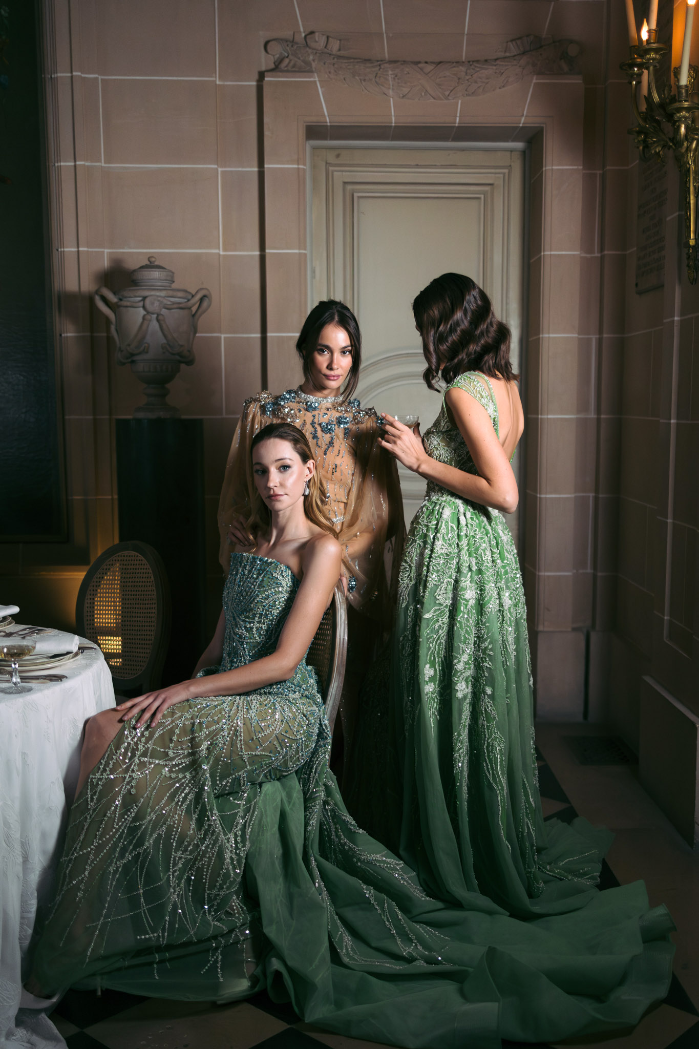 Three women in beaded and embellished gowns posed inside a formal stone-walled interior with candlelit sconces