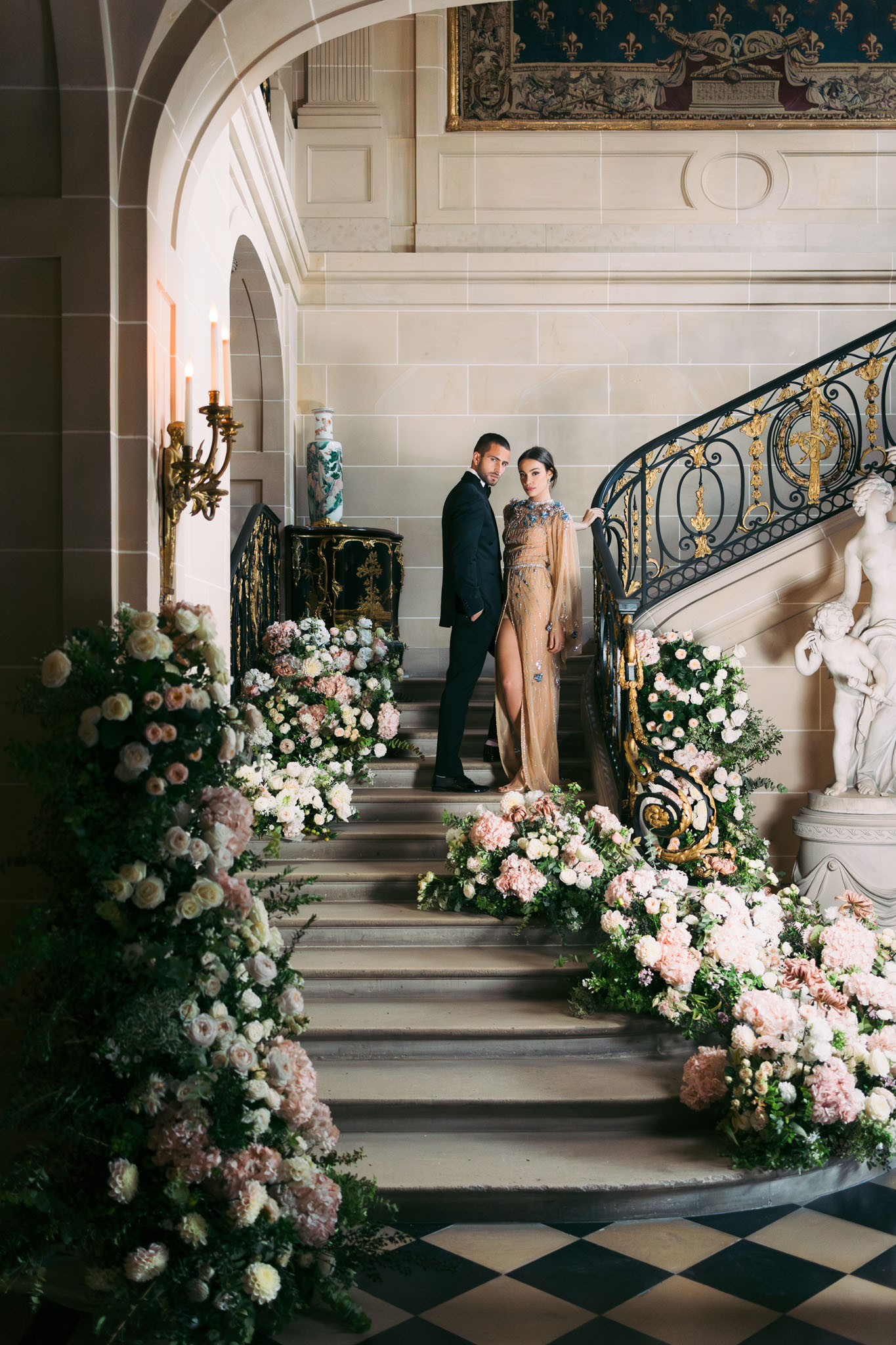 A couple poses together on the grand interior staircase of a French château, standing mid-way up the steps and looking toward the camera. The groom wears a black tuxedo, and the bride wears a floor-length champagne-gold gown with a high slit and embellished neckline featuring blue and silver beading, along with a draped cape detail. The staircase is lined on both sides with large, overflowing floral installations composed of blush pink carnations, dusty rose garden roses, ivory roses, and deep green foliage, cascading from the base of the steps upward. The ironwork banister is black with ornate gold detailing, and a white marble sculptural figure is visible at the base on the right side. The entrance hall features a black-and-white checkered marble floor, stone archways, a gilt wall-mounted candelabra with lit candles, a decorative chinoiserie vase on a lacquered console, and a large tapestry visible at the top of the frame. The shot is a wide portrait taken from the base of the staircase, capturing the full scene. Potential venue feature image.
