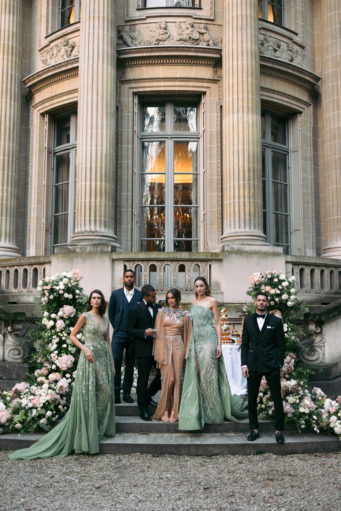 A group portrait of five people posed on the exterior stone steps of a grand French château, featuring ornate Neoclassical architecture with tall pilasters, carved stone friezes, and tall grey-framed windows glowing with warm amber light from inside. Two women wear floor-length sage green embellished gowns with long trains, while a third woman in the center wears a beaded champagne and nude embellished mini dress with a sheer cape. The two men are dressed in dark navy and black tuxedos with bow ties. Flanking the steps are two large floral installations featuring blush pink, dusty rose, and ivory blooms — including roses, hydrangeas, and ranunculus — with lush greenery. A pièce montée (croquembouche) is visible on a table to the right. The overall styling palette is sage green, champagne, and blush against the stone architecture, with a formal, high-fashion aesthetic. Wide editorial-style shot. Potential venue feature image.