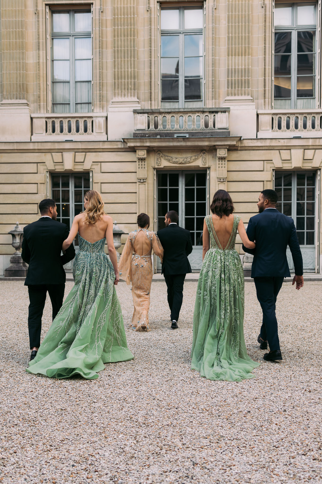 Bridal party of five walking away across gravel courtyard toward grand stone building in sage and gold gowns