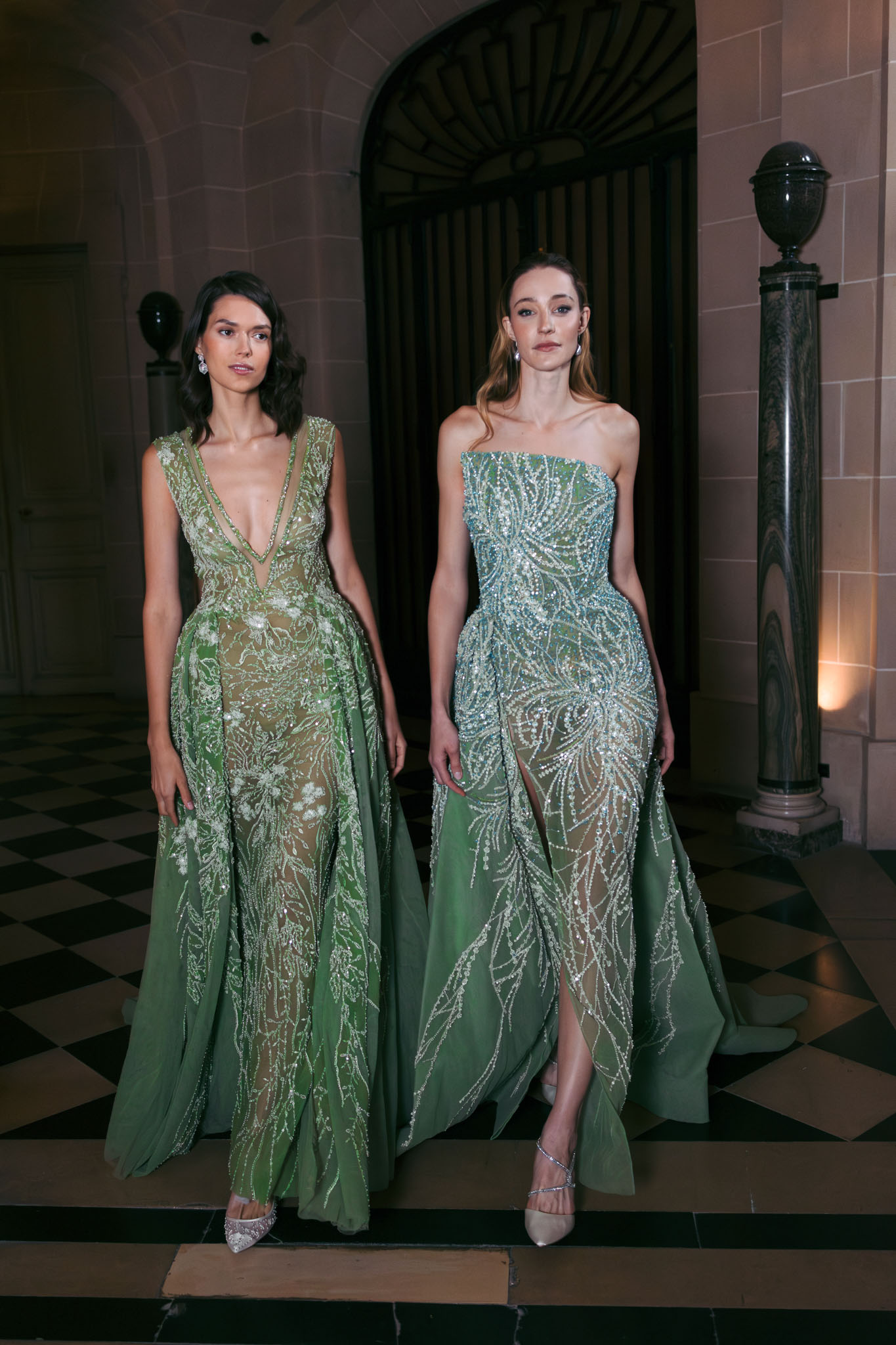 Two women pose together indoors in a grand classical interior featuring a black-and-white checkered marble floor, stone archway, and a dark marble column with warm uplighting. Both women wear floor-length sage and olive green heavily beaded and embroidered gowns with sheer illusion skirt overlays. The woman on the left wears a deep V-neck style with floral beading and a flowing A-line overskirt, while the woman on the right wears a strapless style with crystal and bead embroidery in a swirling pattern and a front slit. Both wear silver strappy heels and drop earrings. The styling is formal and ornate, consistent with a wedding reception or cocktail hour context. Portrait shot, medium distance, with ambient warm lighting from the venue interior.