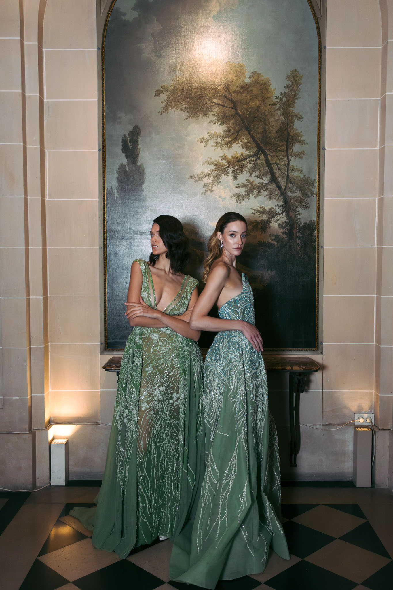 Two women in sage green beaded gowns posing back-to-back in chateau interior with marble floor