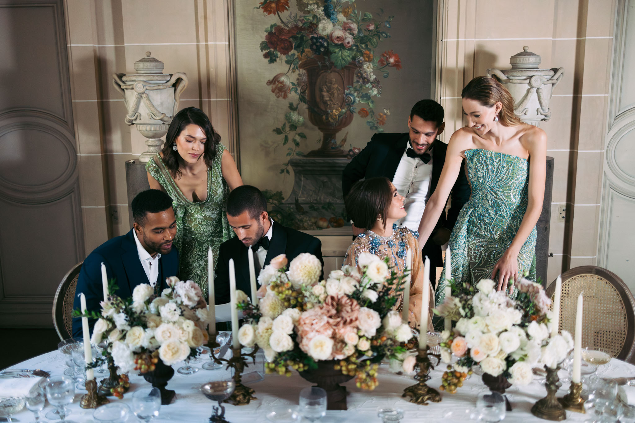 A reception scene shot from a slightly elevated angle, showing six people gathered around a formally set dinner table inside a classical French interior room with painted boiserie panels, ornamental stone urns, and a large oil-style floral mural on the back wall. Two men in black tuxedos are seated at the table, while a third man in a navy suit sits beside them; a woman in a heavily embellished sheer gown with blue crystal details is seated centrally and appears to be in conversation with the standing guests. Two women stand behind the table — one wearing a deep V-neck green sequined gown and the other a strapless teal beaded column gown — both leaning in attentively. The table is dressed in a white linen cloth and features lush low floral centerpieces of ivory garden roses, blush dahlias, mauve blooms, and small berry clusters arranged in dark bronze compote vessels, alongside tall ivory taper candles in ornate gilt candelabras and crystal glassware. The overall styling theme is classic French with jewel-toned fashion and a muted blush and ivory floral palette.