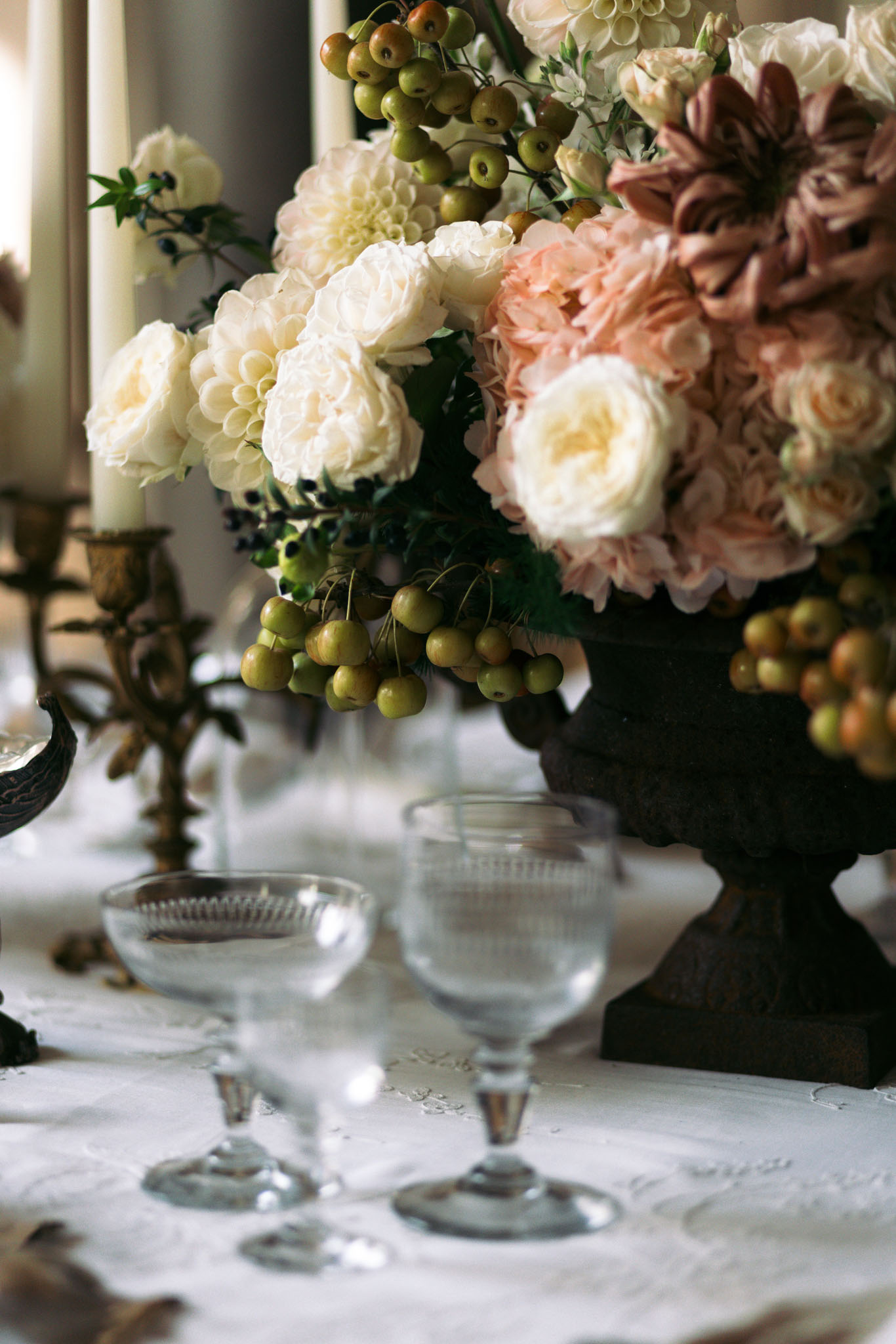 Close-up detail shot of a reception table centerpiece arrangement placed in a dark cast-iron urn. The floral arrangement features ivory garden roses, white and cream dahlias, blush hydrangeas, dusty mauve dahlias, and clusters of small green crabapples as textural accents, with dark berries and dark green foliage woven throughout. In the foreground, two vintage-style cut-glass coupe glasses sit on a white embroidered linen tablecloth, slightly out of focus. To the left, a brass candelabra with unlit ivory taper candles is partially visible. The overall decor palette is ivory, blush, dusty rose, and muted mauve with dark bronze accents, suggesting a classic, richly toned autumn or late-summer aesthetic.
