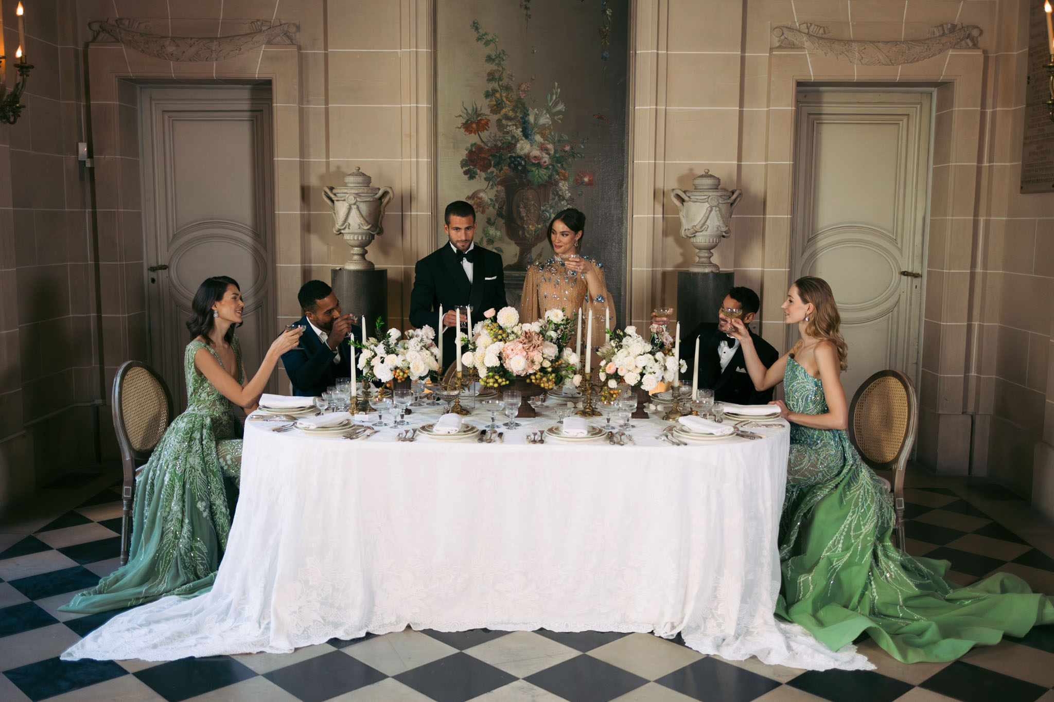 A styled reception tablescape shot taken indoors in a formal château room featuring paneled stone walls, large floral oil paintings, decorative stone urns, wall sconces, and a black-and-white diamond-pattern marble floor. Six people are gathered around a rectangular table covered in a white lace tablecloth: a couple stands at the center — the groom in a black tuxedo and the bride in a champagne-gold long-sleeve gown heavily embellished with blue and silver jeweled embroidery — while two men in dark tuxedos and two women in matching sage green beaded embroidered gowns with long trains are seated on either side, raising glasses in a toast. The table is set with white and gold china, crystal glassware, silver flatware, and folded white napkins; the centerpiece features low arrangements of ivory dahlias, blush garden roses, and cream ranunculus with touches of peach and yellow, flanked by multiple tall ivory taper candles in gold candelabras. The overall styling palette is sage green, champagne, ivory, and gold with a classic, formal aesthetic. Wide shot framing the full table and room symmetrically.